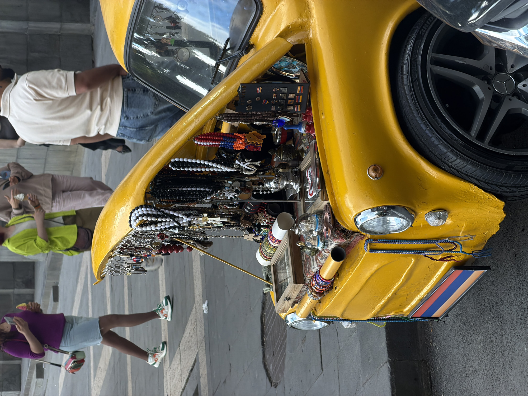 In Yerevan, Armenia, a yellow vehicle's open hood displays an assortment of jewelry for sale. A black tire with a Mercedes logo is visible. Several pedestrians are around the vehicle, including a woman in a purple shirt and light blue shorts.