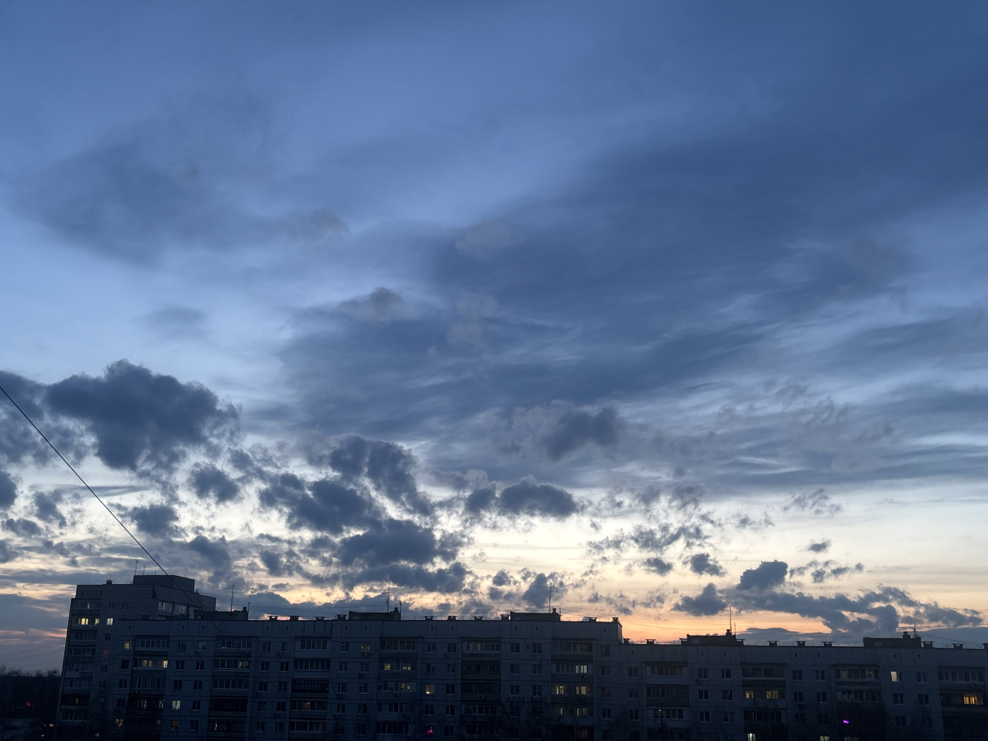 The image displays an urban landscape in Chaikovsky, Russia, during dusk or late evening. The scene is dominated by a vast, cloudy sky and several large, multi-story apartment buildings.
The sky occupies the upper two-thirds of the frame, filled with a dynamic arrangement of clouds. Darker, denser gray clouds are visible in the mid-ground, while lighter, streaky clouds stretch across the upper part, transitioning from deep blue overhead to lighter tones. Towards the horizon, especially on the right, soft warm hues of orange and yellow indicate the setting sun, partially obscured by clouds and buildings.
The lower third of the image features a continuous line of apartment buildings, characteristic of Soviet-era or post-Soviet architecture, silhouetted against the sky. Many windows on various floors are illuminated with artificial light, suggesting that residents are active inside. Faint structures, possibly antennas, are visible on the rooftops. A thin, dark utility wire runs diagonally across the upper left portion of the frame.
No people are visible, and no specific event or activity is taking place beyond the natural transition of day to night. The scene conveys a sense of quiet urban life at twilight. There is no visible text in the image.