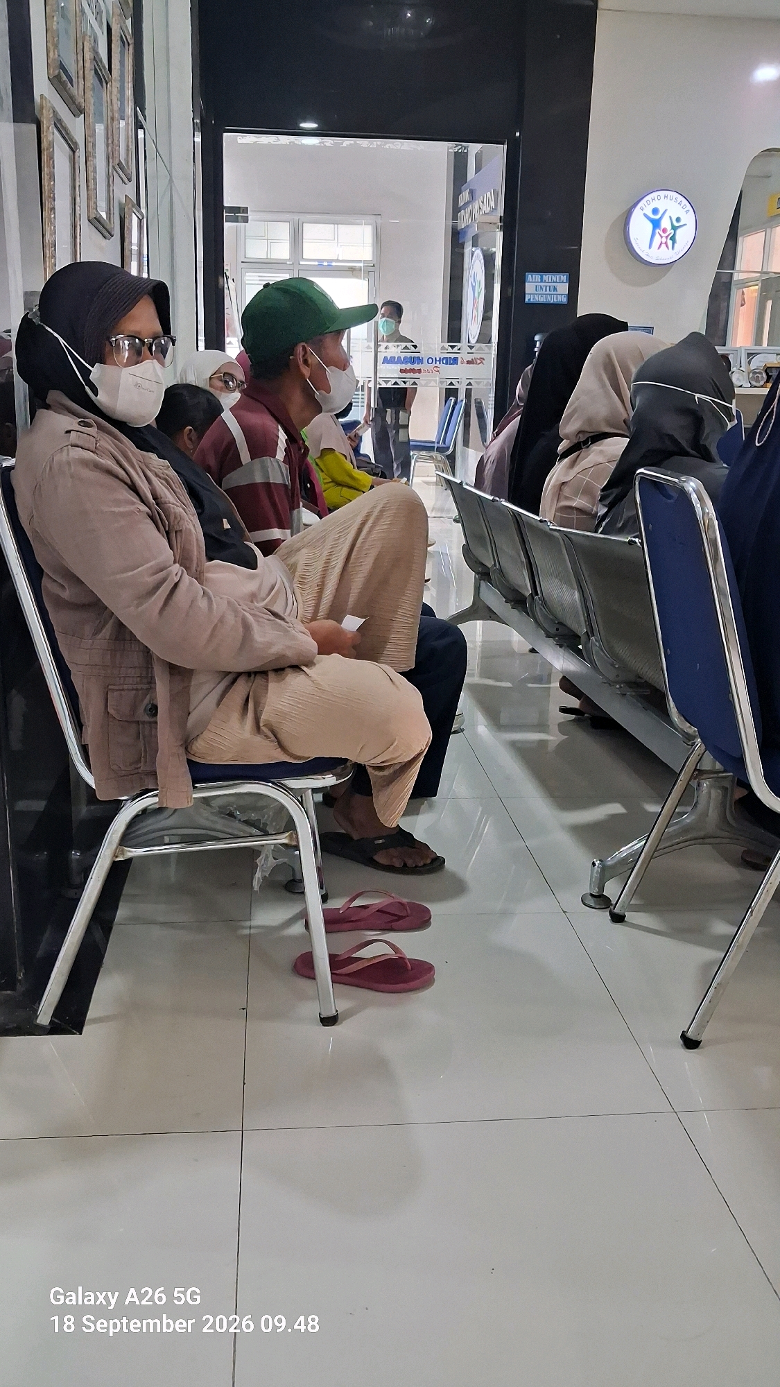 The image shows an indoor waiting area, likely a clinic or hospital, with several people seated on metal chairs. The environment appears to be clean and sterile, with tiled flooring and bright lighting.
In the foreground, a woman wearing a black hijab and glasses is seated, facing slightly to the right. She is wearing a beige jacket and long, light brown pants. Next to her, a person wearing a green cap and a striped shirt is also seated, facing forward. They are wearing a face mask.
Behind them, other individuals are visible sitting on a row of blue and silver chairs. Some are wearing hijabs. Through a glass door in the background, more seating and a person standing can be seen. A circular logo with figures is visible on the wall to the right, and text in Indonesian can be seen on signs.
The overall impression is of a public waiting space, possibly a medical facility, given the presence of face masks and the general clinical atmosphere. The timestamp "Galaxy A26 5G 18 September 2026 09.48" suggests the image was captured on a Samsung Galaxy A26 5G phone on September 18, 2026, at 09:48 AM. The mention of Kutoarjo, Indonesia, would indicate the location of this waiting area.