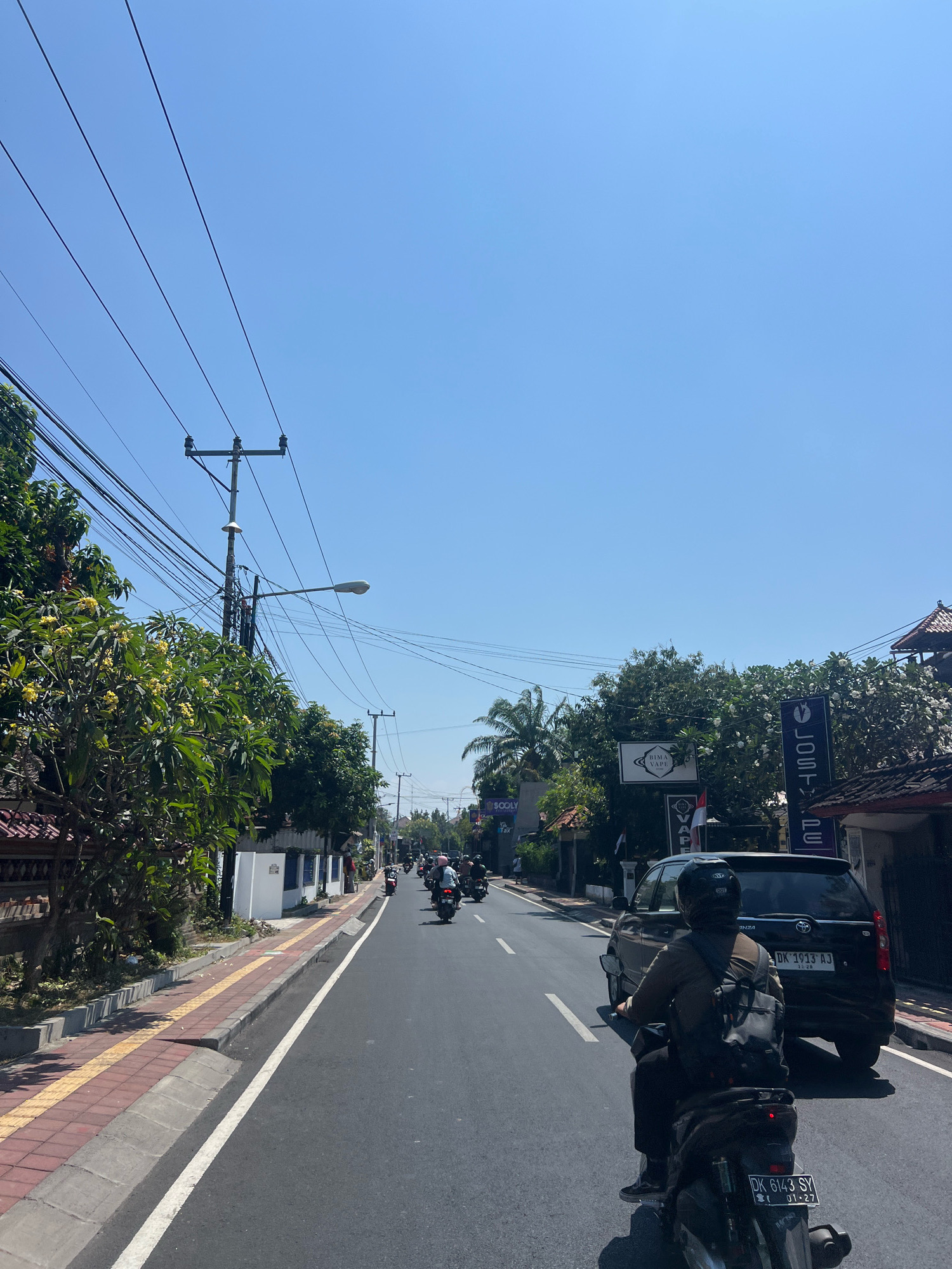 This is a photograph taken from a motorcycle on a sunny day in Denpasar, Indonesia. The road is paved with asphalt and marked with white lines. Multiple motorcycles are visible on the road, with riders wearing helmets. A black SUV is also present. On the left side of the road, there is a sidewalk paved with red bricks and yellow tiles, bordered by a curb. Trees and foliage line the sidewalk, and a power pole with numerous wires is prominent. Several signs are visible on the right side of the road, including one that says "LOST VE DE" and another for "BAMA VAPE". The sky is clear and blue.