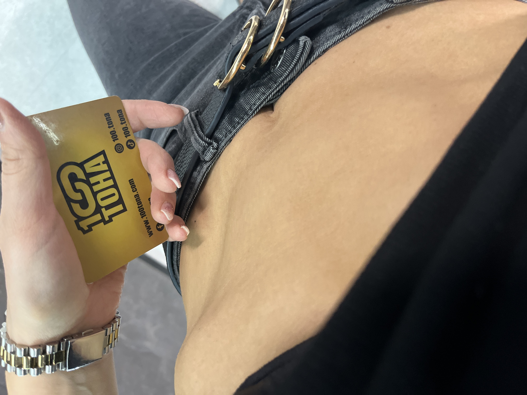 The image shows a person holding a gold-colored card in their left hand. The card is branded with "100 TONA" and features the website address "www.100tona.com" along with social media icons for Instagram and TikTok. The person is wearing grey jeans with a gold-colored double-buckle belt and a black top. A two-toned gold and silver wristwatch is visible on their left wrist. The image does not provide enough information to determine the specific location in Varna, Bulgaria.