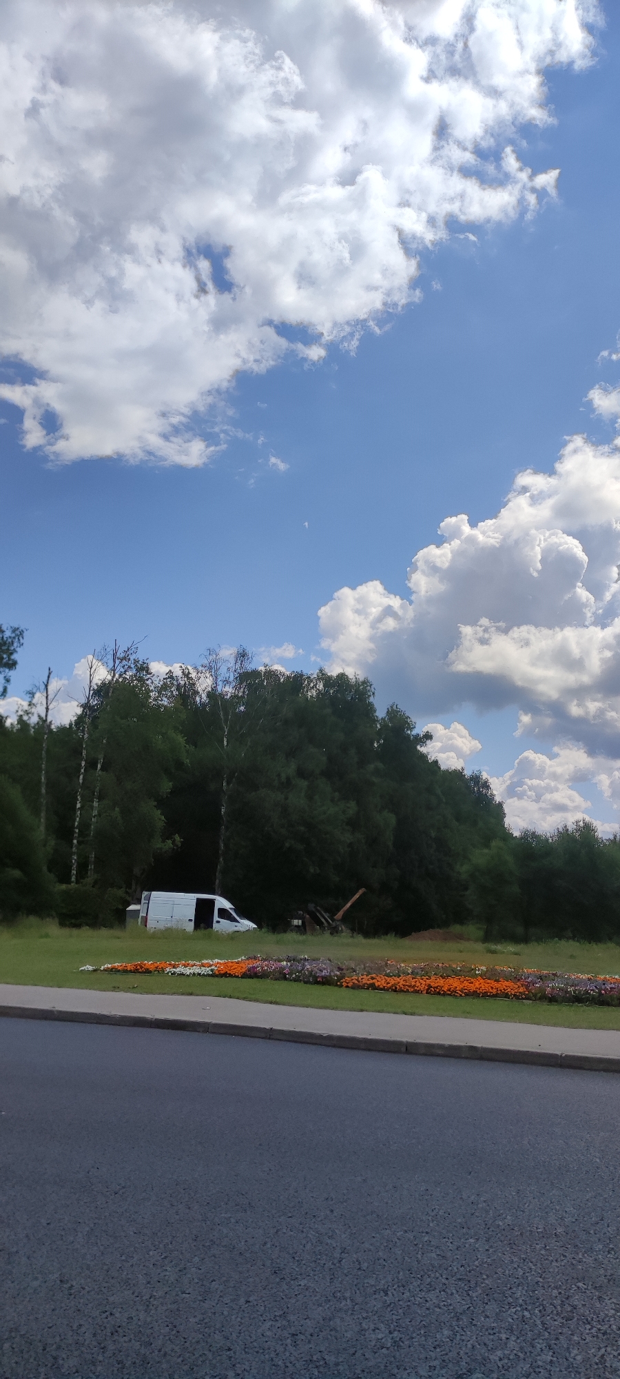 The image captures a bright, cloudy day in Vatutinki, Russia, with a clear blue sky dominating the upper half. A white van is parked on a grassy area, its side door open. In front of the van, a long, colorful flowerbed stretches horizontally, featuring rows of orange, white, and purple blooms. Behind the van and flowerbed, a dense line of trees forms a dark green backdrop. To the right of the van, a piece of orange construction equipment is visible amongst the trees. The foreground shows a paved road with a gray curb.