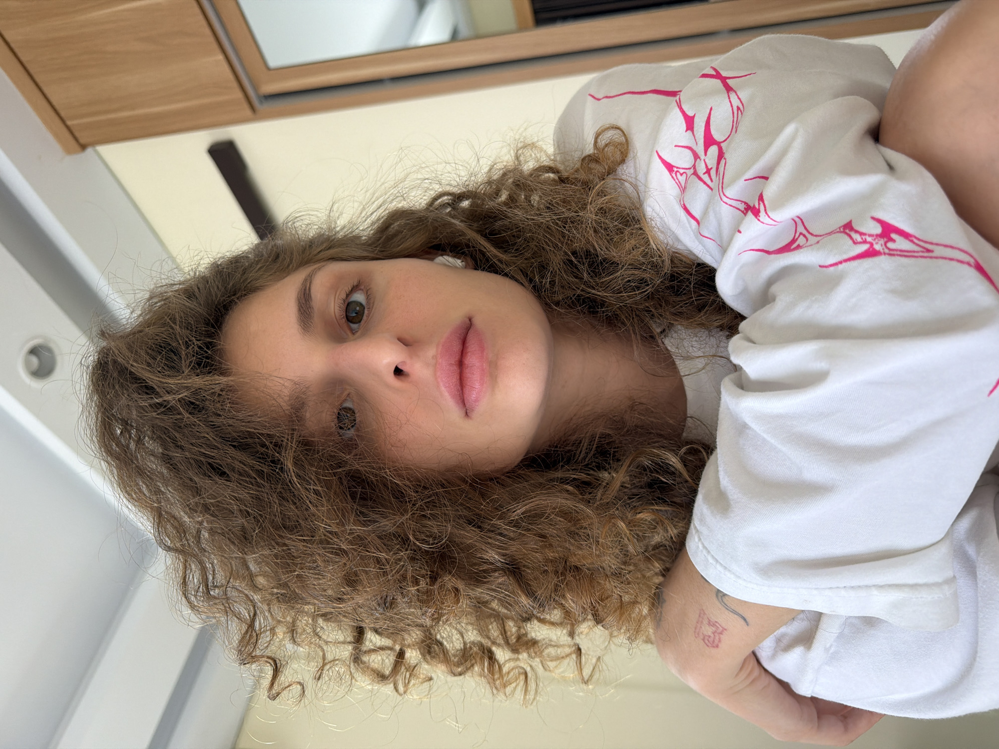 A young woman is depicted in a close-up, upward-angle portrait, reportedly taken in North Kuta, Indonesia. She has voluminous, medium-brown curly hair that frames her face and extends beyond her shoulders. Her expression is neutral, with her gaze directed precisely at the camera. Her eyes are brown, and her lips are tinted with a soft pink. She wears a white long-sleeve t-shirt featuring distinct fuchsia-pink graphic designs, resembling abstract flame or tribal motifs, primarily visible on the sleeves. A small white wireless earbud is present in her right ear. On her left inner wrist, two small tattoos are visible: one appears to be a red/pink stylized symbol or character, and the other a thin black line. Her arms are partially visible, crossed or resting in the lower foreground. The background reveals an indoor setting with light-colored walls, a white ceiling with a recessed circular light fixture, and a section of a light-toned wooden cabinet or wardrobe with a mirror on the right side. A dark, horizontal structural element is also visible on the upper right wall.
