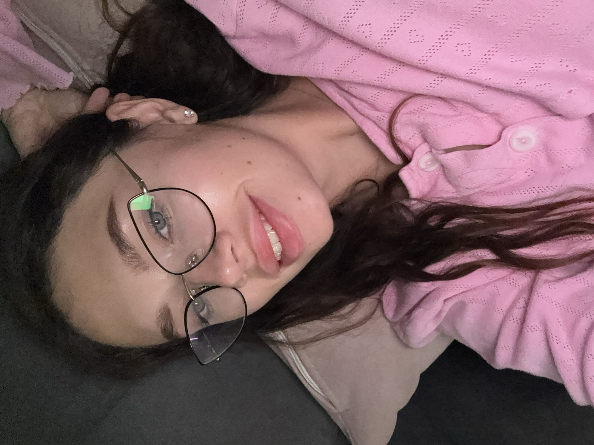 The image captures a young woman with long, dark brown, wavy hair, lying down on what appears to be a dark grey or black couch or bed. Her head is tilted, giving the photograph a rotated perspective. She is wearing round, wire-rimmed glasses, with a light reflection visible on the left lens. Her blue-grey eyes are directed towards the camera, and she displays a soft, pleasant smile, with her lips slightly parted to reveal her upper teeth. Her skin appears clear, and her eyebrows are well-defined.
She is dressed in a light pink, long-sleeved top or cardigan, distinguished by a delicate openwork knit pattern throughout the fabric. The garment features white buttons on the front. A small, sparkling stud earring is visible in her left ear. Her hair is spread around her head and shoulders, with some strands covered by the pink clothing.
Her head rests on a light-colored, possibly beige or off-white, pillow. The background consists of the dark grey/black surface and more of the pink fabric, suggesting a comfortable, indoor setting. The lighting is soft and even, indicating either indoor lighting or diffused natural light. The overall impression is one of relaxation or a casual selfie taken in a home environment in Tel-Aviv, Israel. No visible text is present in the image.