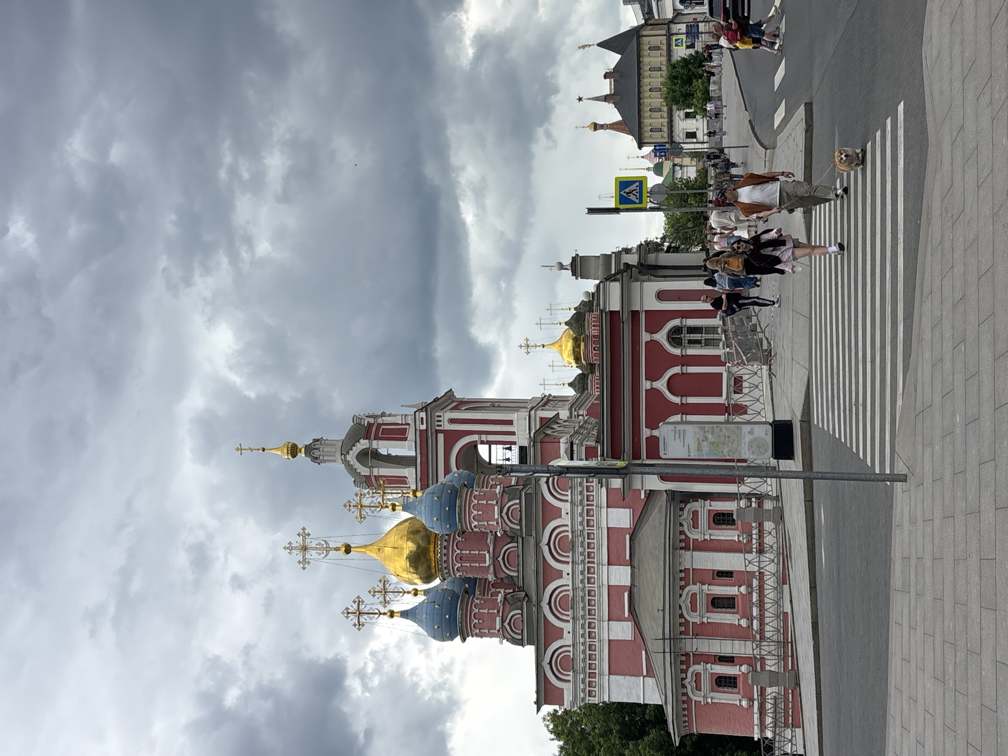 The image shows a building with red and white walls, a church or cathedral in Moscow, Russia, with gold and blue domes topped with crosses. The sky is overcast and cloudy. People are walking across a pedestrian crosswalk in the foreground, including a person walking a small dog. In the background, other buildings can be seen.