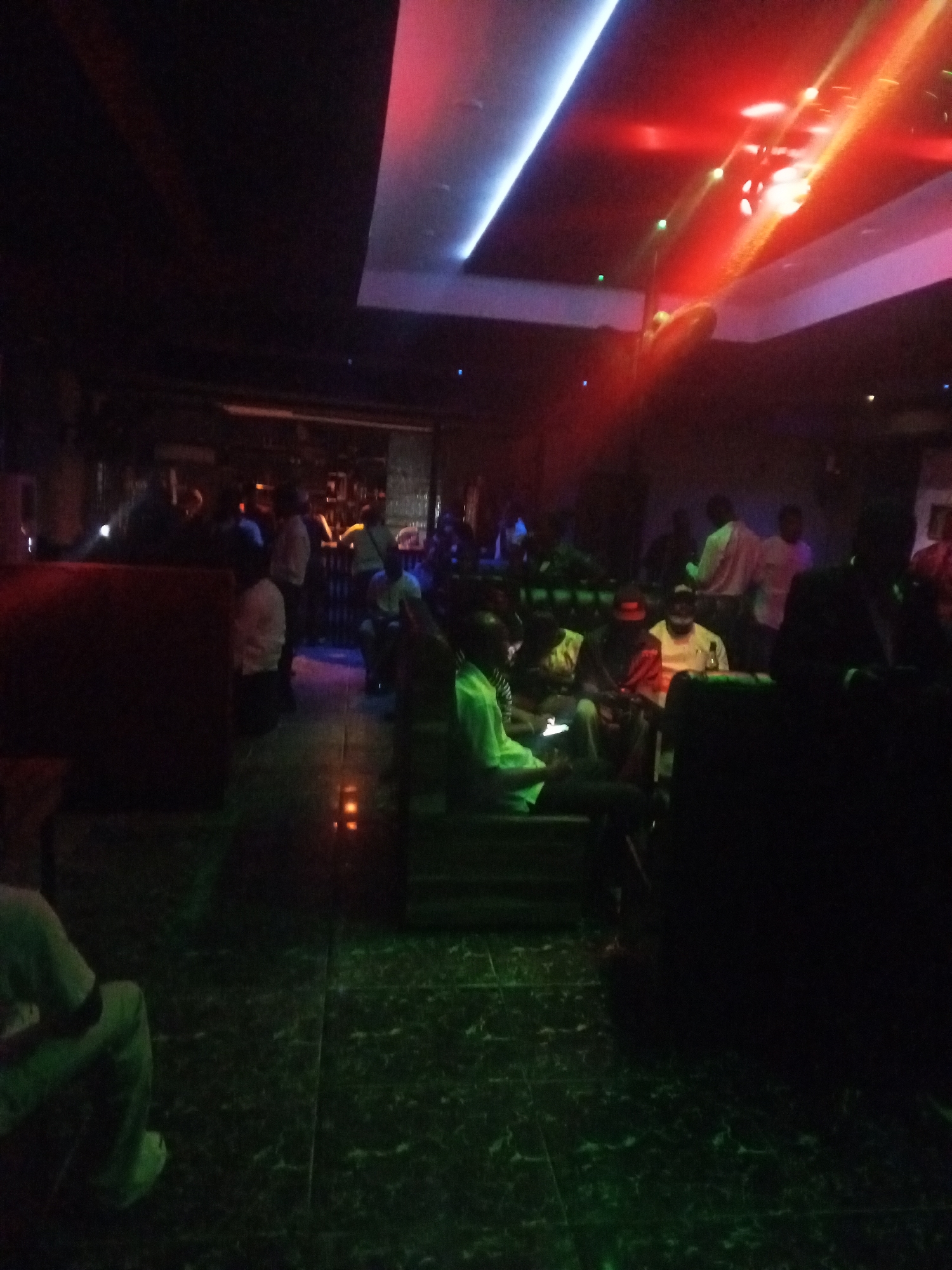 The image depicts an indoor scene, likely a nightclub or bar, with dim lighting and colored spotlights. Several individuals are visible, some seated and others standing. A bar area with bottles is discernible in the background. The floor appears to be tiled with a green reflective surface. Red and blue lights illuminate the ceiling. The overall atmosphere suggests a social gathering in a dimly lit establishment.
