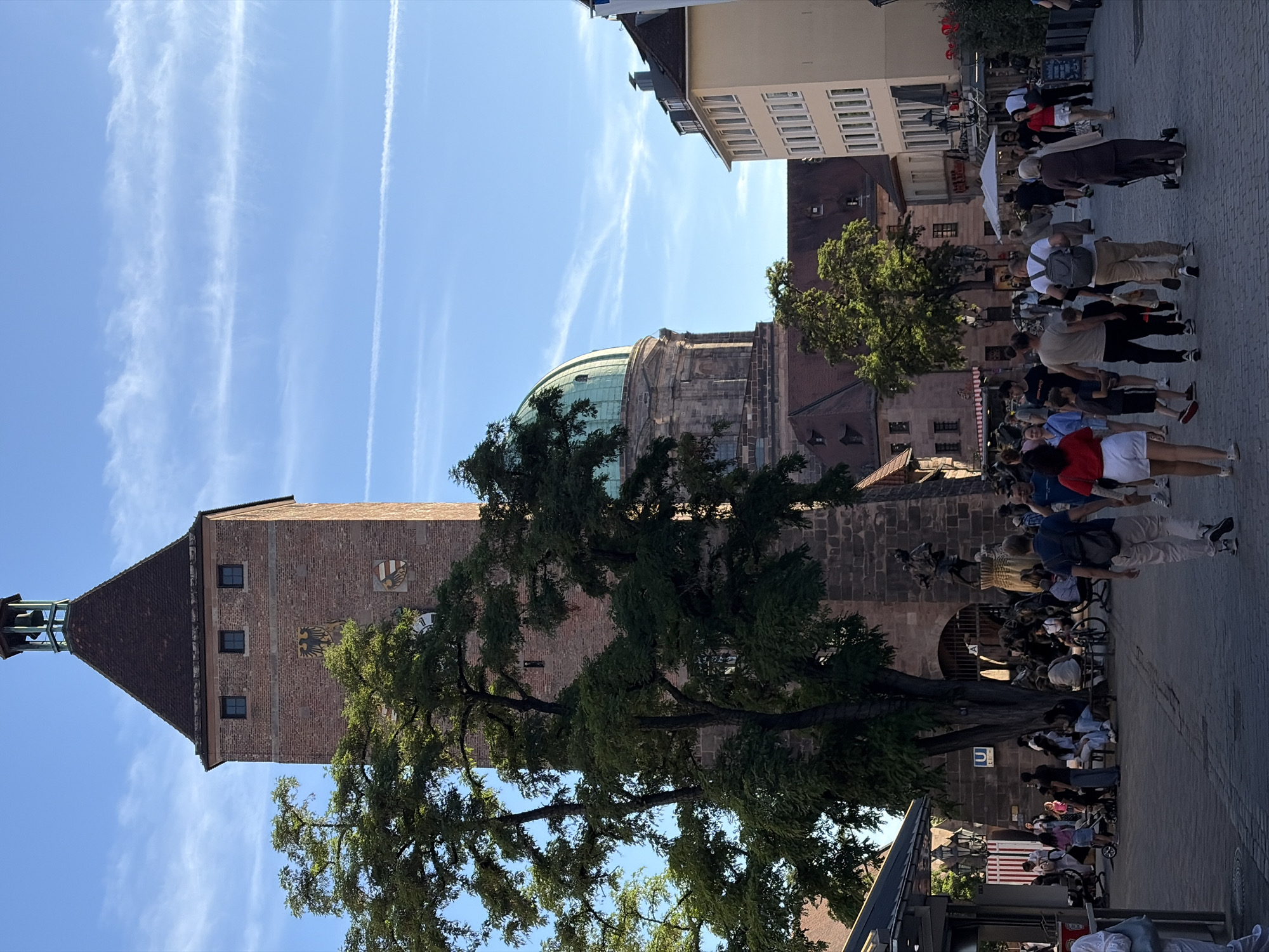 The image captures a bustling scene in Nuremberg, Germany, likely near the historic Handwerkerhof or a similar marketplace area adjacent to the medieval city walls. A prominent red-brick tower with a dark tiled, pyramidal roof dominates the left side of the frame, topped with a small bell. The tower features small, dark windows and two coats of arms. Behind the tower, a large, green dome of a building is visible.
In the foreground, a paved pedestrian area is filled with people walking and gathering. They are dressed in casual summer clothing. Several individuals are walking towards the viewer, while others are seen from behind or the side. Some people are sitting on benches or near a fountain structure at the base of the tower. A large tree with dense green foliage stands in front of the tower, partially obscuring it.
The background shows several multi-story buildings with light-colored facades and windows, typical of European city architecture. White umbrellas suggest outdoor seating or market stalls. The sky is a clear blue with wispy white clouds and contrails from airplanes, indicating fair weather. The overall impression is of a lively public space on a sunny day, with historical architecture serving as a backdrop to daily life and tourism.