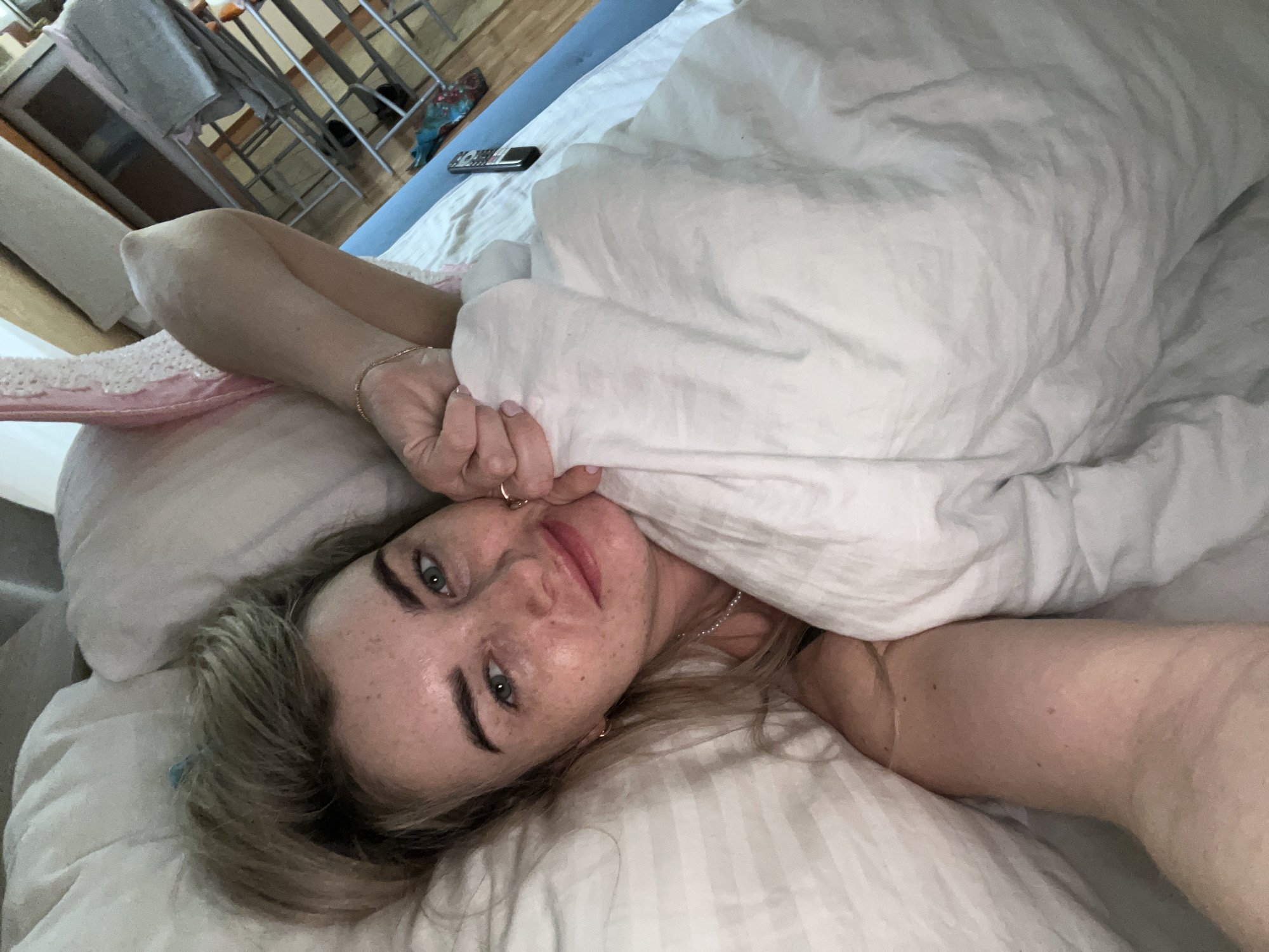 An intimate, high-angle shot captures a woman lying in bed, looking directly at the camera. She is positioned on her back, with her head resting on a white striped pillow and her body partially covered by a white duvet or blanket. Her light-colored hair, possibly blonde or light brown, frames her face, which is adorned with freckles. She has blue or green eyes and a relaxed expression, with a subtle hint of a smile. She wears small hoop earrings and a delicate pearl necklace. One of her hands is visible near her face, appearing to grasp the duvet.
The bed features white bedding, including the duvet and pillow, and a blue fitted sheet is visible beneath the white layers. A black remote control rests on the bed near the headboard. The background is blurred but reveals elements of an indoor room, including what looks like a clothes rack or shelving unit and a light-colored floor, suggesting a bedroom setting. The lighting is soft and diffused, characteristic of an indoor environment without strong direct sunlight, possibly early morning or daytime. No explicit text is visible within the image. The scene conveys a sense of comfort and repose, set in Chaikovsky, Russia.