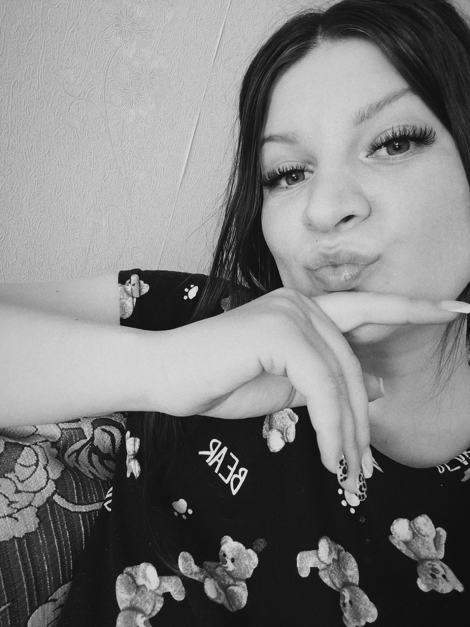 A monochrome, medium-shot photograph depicts a woman with long, dark hair and full eyelashes. Her facial expression is one of puckered lips, and her hand is positioned with the index finger extended to touch her chin. Her nails are long and painted with a leopard print design. She is wearing a dark-colored top with a repeating pattern of teddy bears. The background is a textured wall. Part of a patterned armchair can be seen. There is no information that suggests this photograph was taken in Tobolsk, Russia.
