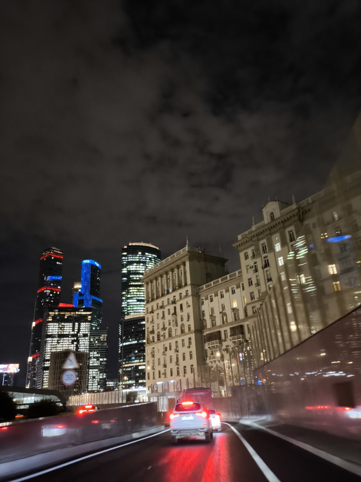 In an urban nighttime scene, a multi-lane road is visible, with cars in motion. The road surface appears wet, reflecting the lights of vehicles and buildings. To the left, a row of modern skyscrapers with illuminated facades, featuring red and blue lighting on some, dominates the background. One skyscraper displays a large digital clock. Adjacent to the skyscrapers, an older, multi-story building with numerous windows is present. To the right, another tall building, possibly a skyscraper, is partially visible, its glass facade reflecting city lights. The sky is dark and cloudy. The perspective suggests the image was taken from inside a moving vehicle.