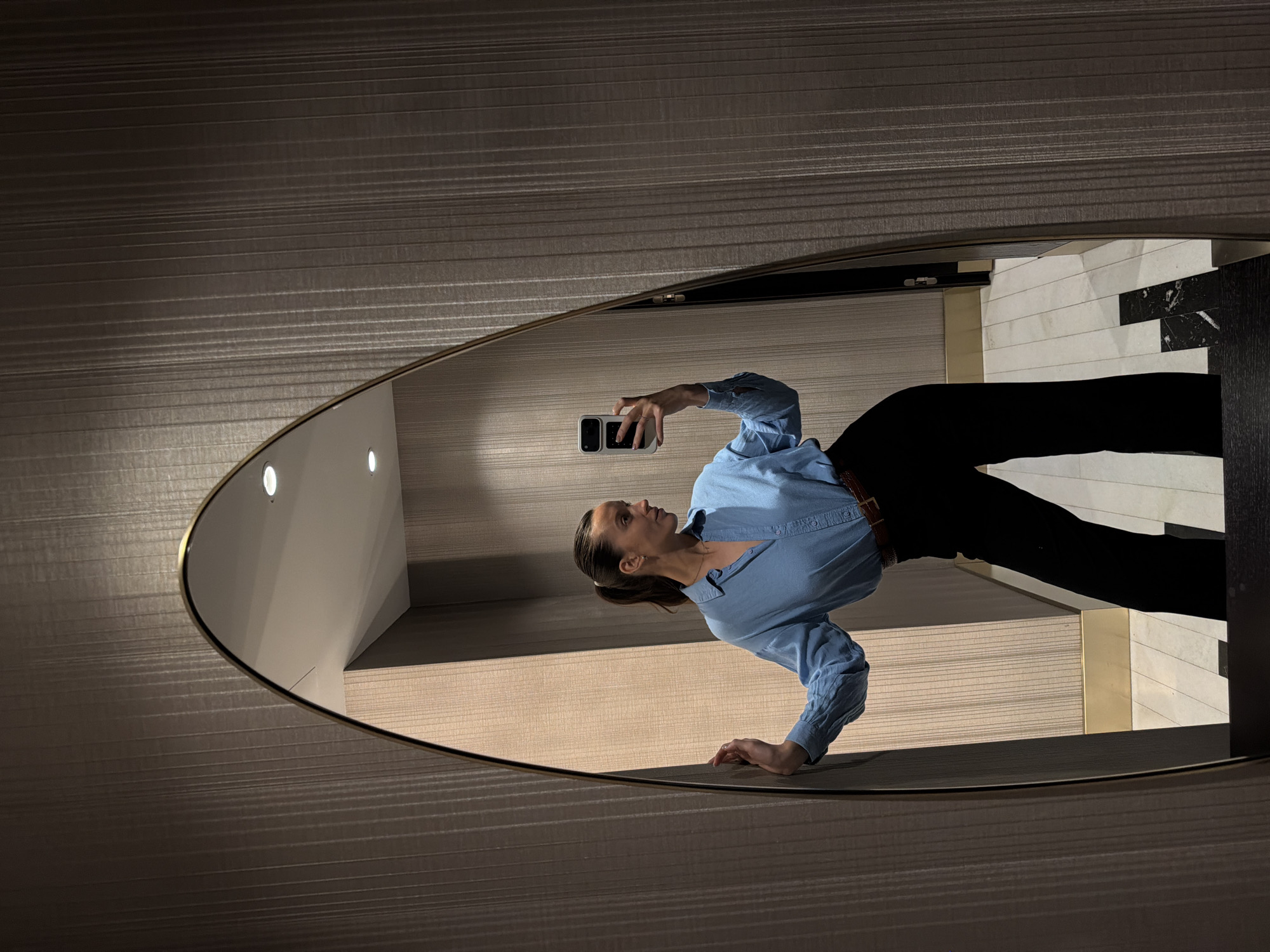 An indoor mirror selfie captures a woman taking her picture using a smartphone. The woman is visible from the torso up in the reflection, standing slightly to her left and looking upwards towards the phone she holds with her right hand. She is dressed in a light blue long-sleeved button-up shirt, black trousers, and a brown belt, with her hair pulled back.
The reflection is contained within a large, irregularly shaped mirror, distinguished by a subtle golden-toned frame. The immediate environment surrounding the mirror features textured walls with horizontal stripes in muted brown and grey tones.
Within the mirror's reflection, behind the woman, elements of a contemporary room are visible. These include a wall clad in white tiles with intermittent black marble sections, and a dark-toned counter or vanity. Several circular downlights are also reflected above this surface. The overall lighting is soft and artificial, characteristic of an interior space. There is no visible text in the image. The setting for this scene is Budva, Montenegro.