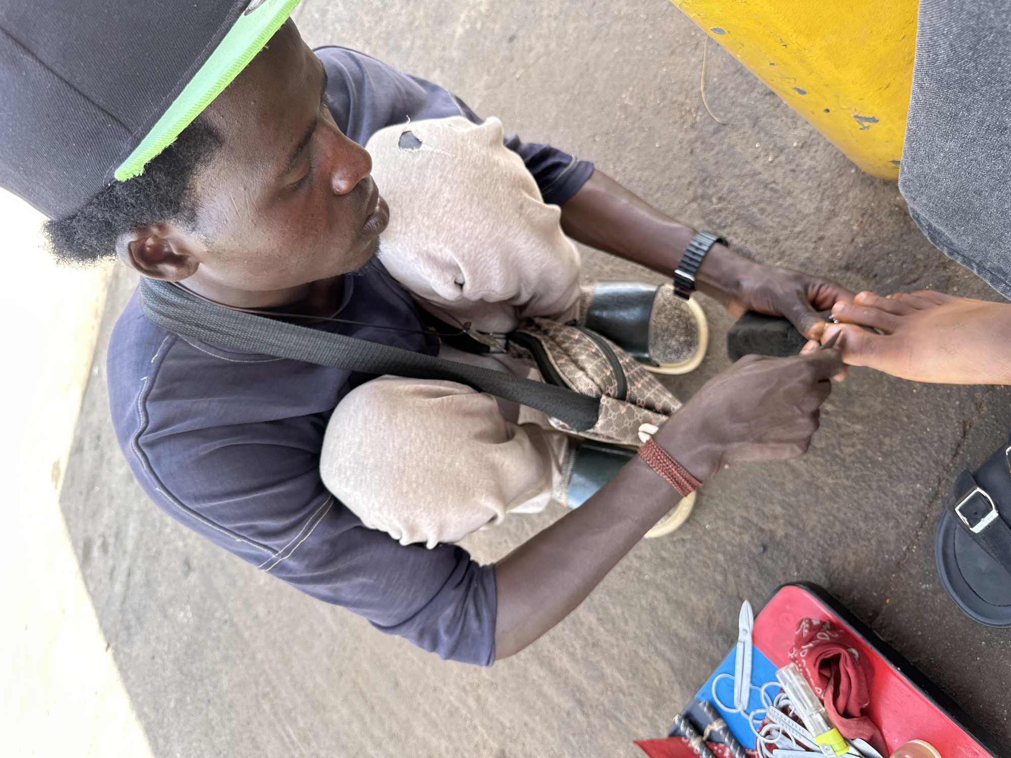 A high-angle shot captures a dark-skinned man providing a pedicure service in an outdoor setting, likely in Jos, Nigeria. The man, appearing to be seated or squatting, wears a black baseball cap with a bright neon green brim, a dark blue short-sleeved shirt, and dark trousers. His short, curly dark hair is visible under the cap. He has a black wristwatch on his left wrist and a reddish-brown braided bracelet on his right.
Resting on his lap is a light-colored, possibly off-white, folded garment with a small tear, partially obscuring a patterned shoulder bag. He is intently focused on the bare foot of another person, visible on the right, holding it with one hand and using a small, metallic grooming tool on the toes with the other. The person whose foot is being groomed wears a dark sandal with a silver buckle.
On the ground next to the service provider, a small, red and blue tray holds various tools, including scissors, nail clippers, and other small instruments, along with a piece of red fabric. The ground is a rough, light gray concrete or paved surface. A bright yellow painted structure is visible in the upper right background. The natural, bright lighting indicates it is daytime. The man's expression is concentrated on his task. No text is visible in the image.