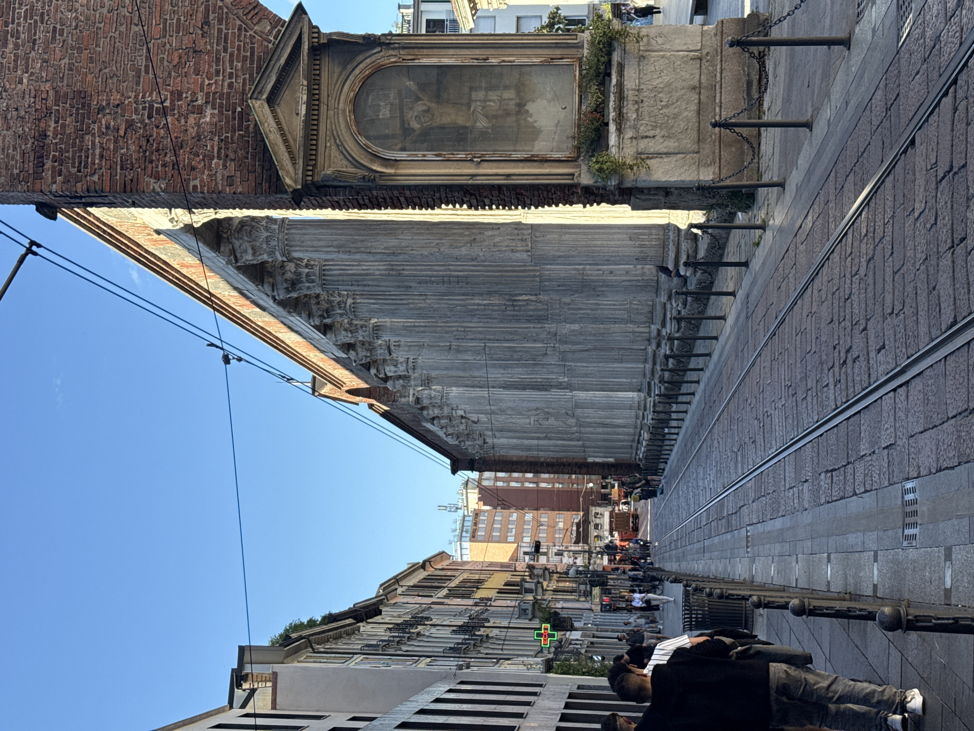 The image captures a street view in Milan, Italy. The subject's vantage point is located on a sidewalk, with a row of columns visible on the left side, leading towards the background. Above the columns is a brick structure with an arched window.
To the right of the sidewalk are tram tracks embedded in a cobblestone street, running parallel with the columns and the sidewalk. A chain fence with metal posts is visible along the edge of the sidewalk, separating it from the street.
Pedestrians can be seen walking along the street, and buildings line the street in the background. The sky above the buildings is clear blue.
The orientation of the photograph is rotated ninety degrees.