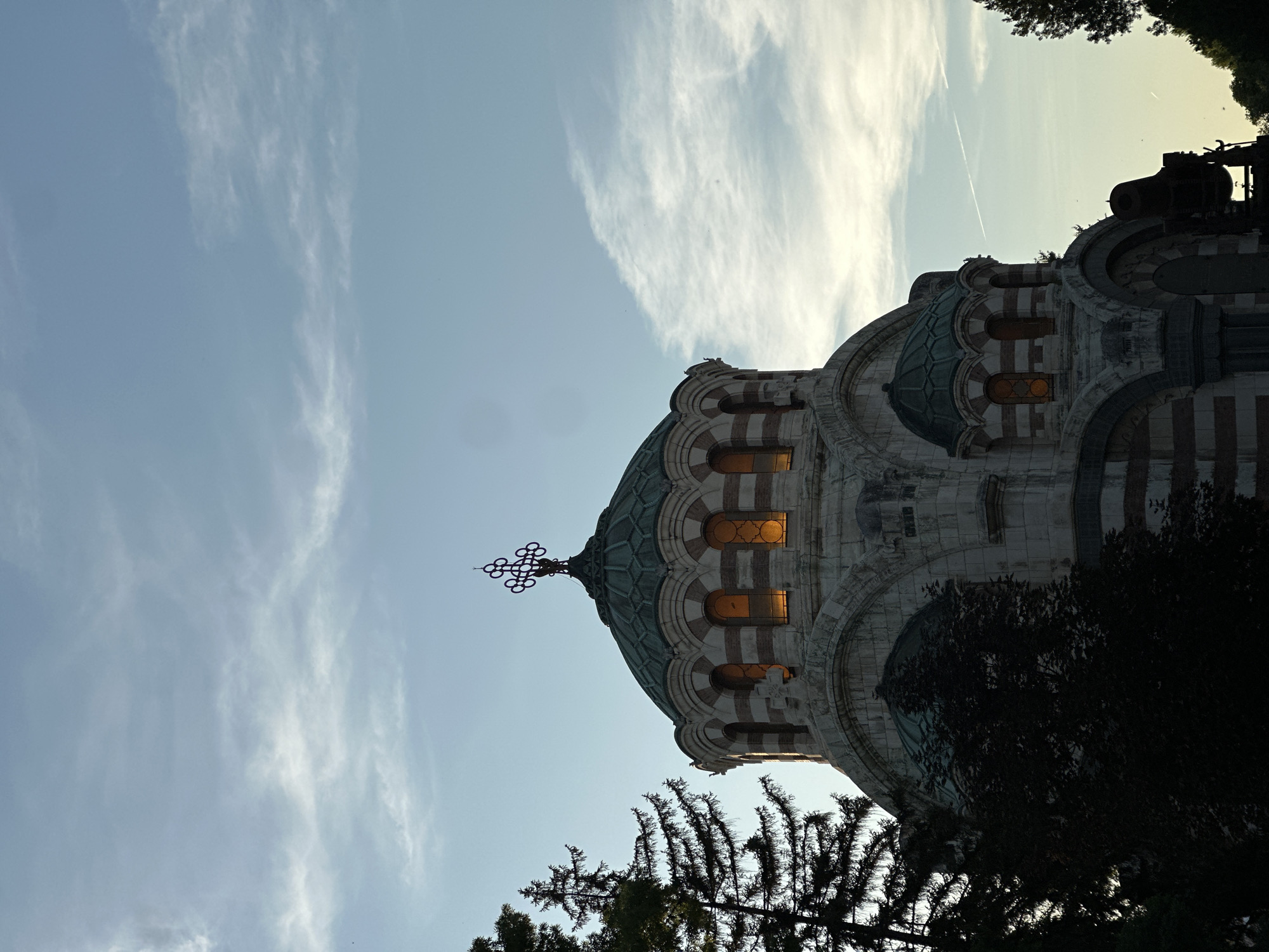 The image shows the Pleven Panorama, a memorial complex in Pleven, Bulgaria, commemorating the Russian-Turkish War of 1877-1878. The foreground features a large, domed building with arched windows that are lit from within, suggesting it is dusk or evening. The architecture is reminiscent of traditional Orthodox churches, with a large central dome and smaller domes, adorned with a distinctive cross on top. The building is constructed of stone and features striped bands of red and white. To the left of the building, the dark silhouette of a tall pine tree is visible, its branches reaching towards the sky. In the lower right portion of the frame, the dark silhouette of what appears to be an old cannon on a mount is discernible. The background consists of a pale blue sky with wispy white clouds, and a faint contrail from an aircraft. The overall impression is one of historical significance and solemnity, set against a natural backdrop.