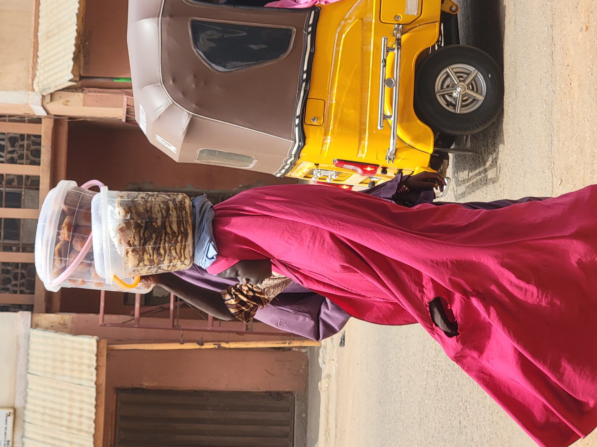 A woman in a bright pink and purple dress is walking down a street in Maiburiji, Nigeria, carrying two transparent plastic containers of food on her head. Behind her is a yellow auto rickshaw. The background includes buildings with metal grates and corrugated iron roofs.