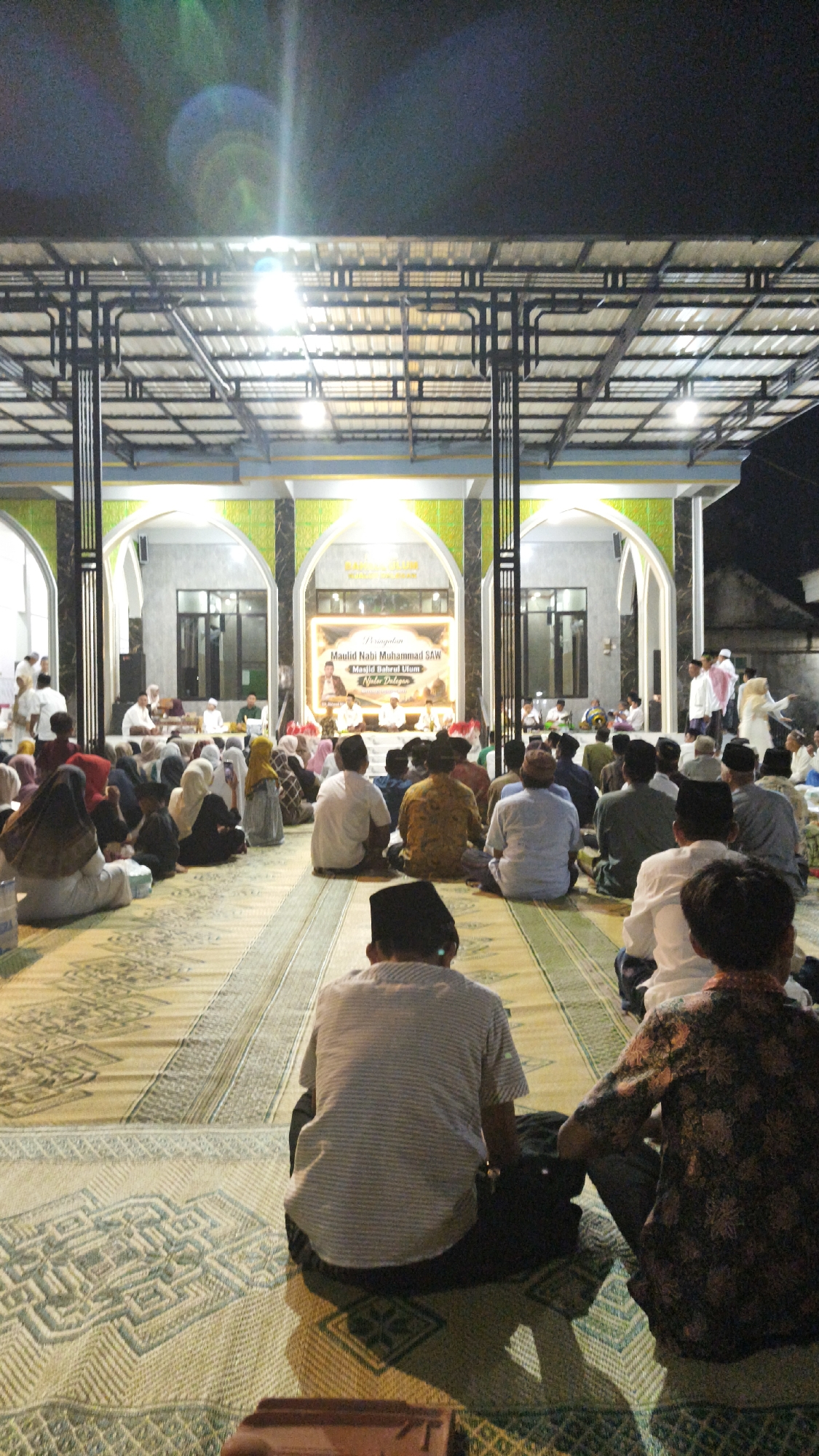 The image depicts an evening gathering at the Masjid Bahrul Ulum in Warulor, Indonesia, celebrating Maulid Nabi Muhammad SAW. A large group of people are seated on the floor, facing a stage area where several individuals are present. The mosque's architecture is visible, with arched entrances and a metal roof structure illuminated by artificial lights. The floor is covered with woven mats displaying intricate patterns. A banner on the stage reads "Peringatan Maulid Nabi Muhammad SAW Masjid Bahrul Ulum" and includes images of religious figures. The atmosphere appears to be one of religious observance and community gathering.