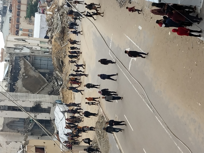The image displays a street scene in Khan Yunis, Palestinian Territory, characterized by extensive urban destruction and human activity. In the background, several multi-story buildings are severely damaged or partially collapsed, exhibiting exposed rebar, shattered windows, and significant structural compromise. Large piles of concrete rubble and debris are scattered across the ground in front of these structures. Two large white tents are visible in the mid-ground, situated among the debris and damaged buildings.
Approximately 50 to 70 individuals, a mix of adults and some younger persons, are dispersed throughout the scene. They are seen walking along a partially paved road, which features worn white lane markings, and across unpaved, dusty areas near the rubble. Most individuals are dressed in casual, dark-colored clothing. A thick black cable or hose lies along the edge of the paved road. The sky is overcast. The scene reflects a populated area significantly impacted by damage, with people moving through the environment.