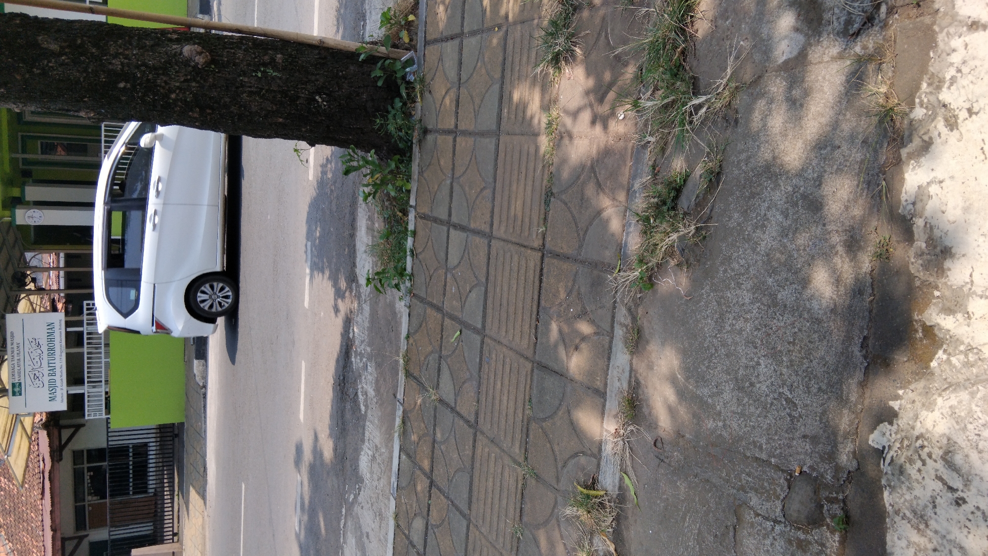 In Proyonangg, Indonesia, a white minivan is parked on the side of a paved road in front of a mosque. The mosque, named Masjid Baiturrohman, has a green wall and a sign with Arabic and Indonesian writing. A large tree with dark bark stands next to the sidewalk, casting shadows on the ground. The sidewalk is made of concrete tiles with decorative patterns and some grass growing between them. The road is asphalt and appears to be a street.