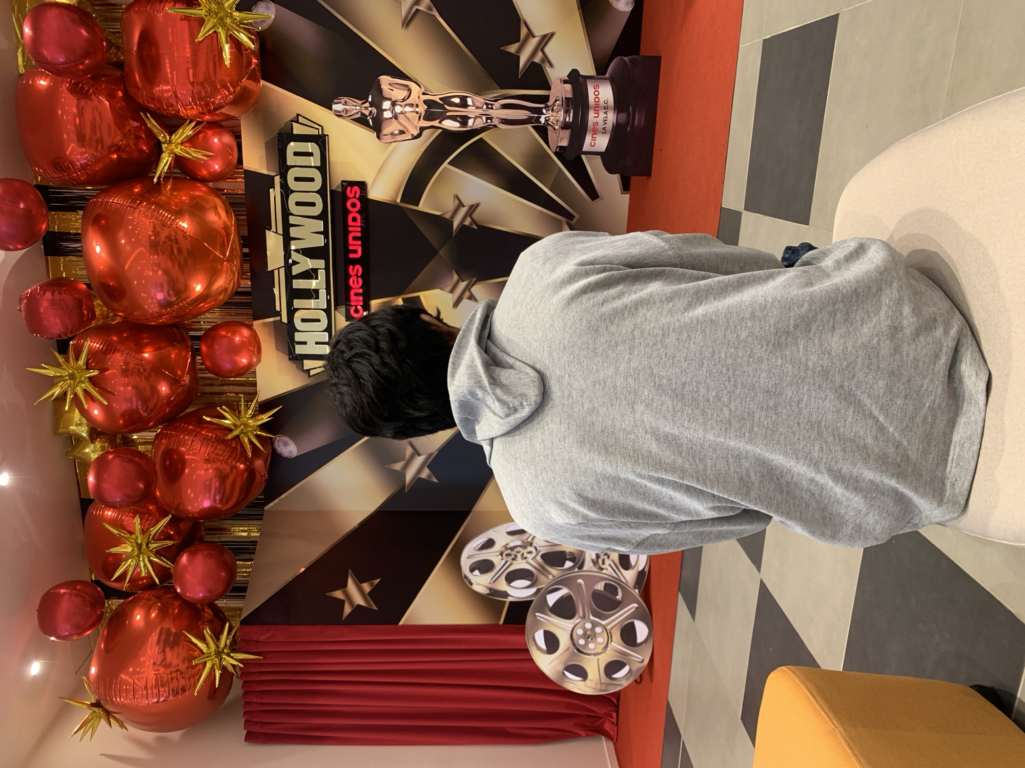 A person wearing a gray hooded sweatshirt sits with their back to the camera in a decorated room. The decorations include red balloons with gold stars, a backdrop featuring "HOLLYWOOD" and "CINES UNIDOS" signage, a golden Oscar statue image, and film reel cutouts. The flooring has a geometric pattern of alternating black and white shapes. A red curtain is present. An orange chair is also visible. The scene appears to be set up for a promotional event or gathering related to movies, possibly at the location of Cines Unidos La Vela in Porlamar, Venezuela.