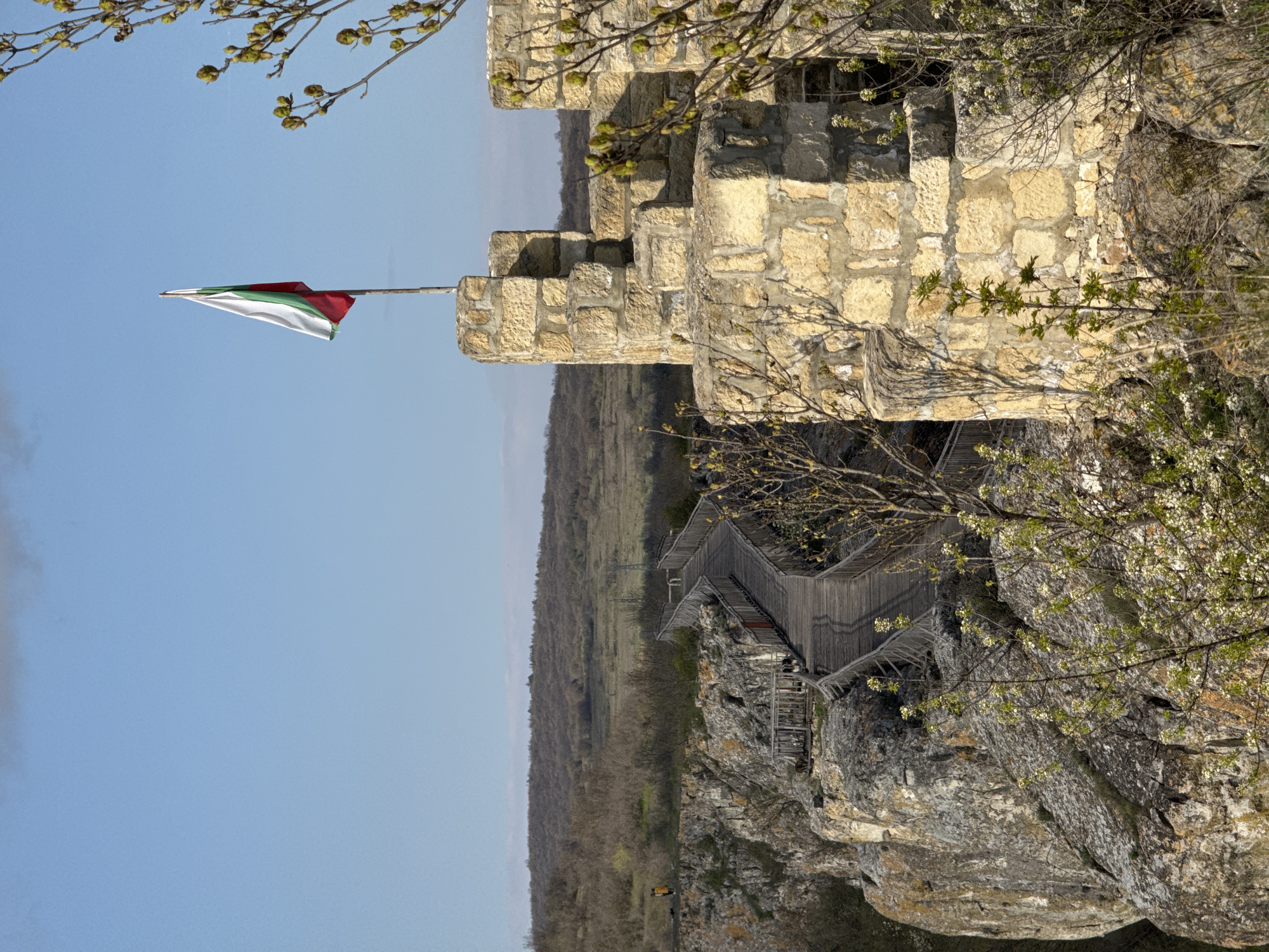 The image shows the Ovech Fortress, located in Provadia, Bulgaria. A stone tower with a flag (white, green, and red) stands atop a cliff overlooking a valley. A wooden walkway and stairs descend the cliff face. Vegetation is present on the cliff and in the background. The sky is light blue.