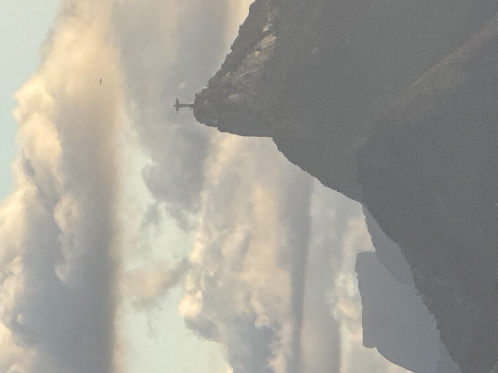 The image shows a view of the Christ the Redeemer statue atop Corcovado Mountain, overlooking the city of Rio de Janeiro, Brazil. The statue is silhouetted against a cloudy sky. In the distance, other mountains can be seen, including what appears to be the city of Niterói across Guanabara Bay. A small aircraft is visible in the sky to the right of the statue. The overall lighting suggests either early morning or late afternoon.