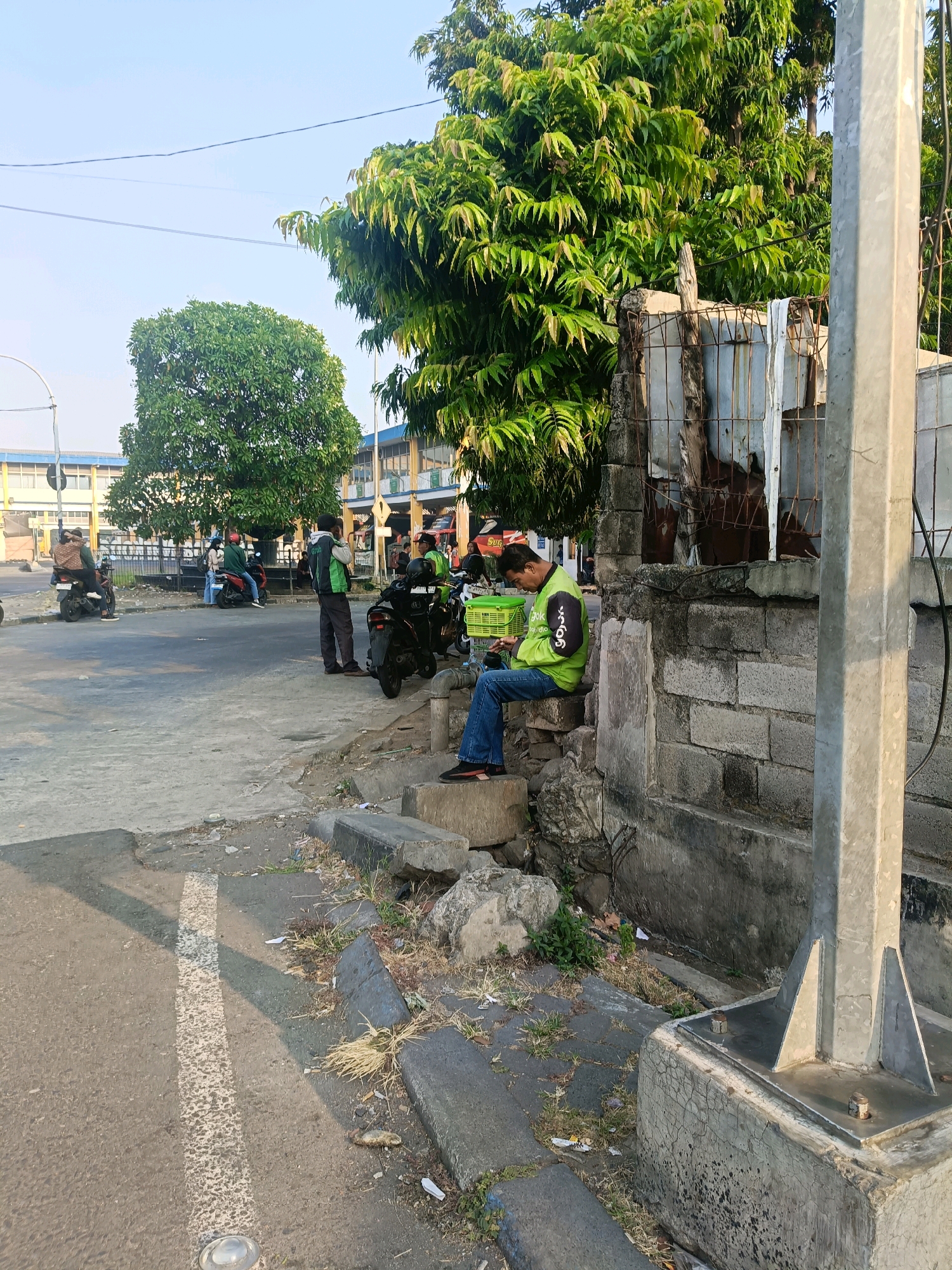 In Kedungrejo, Indonesia, on a paved road with a white line marking the edge of the lane, a man in a lime green jacket and blue jeans sits on a low concrete barrier, looking at his phone. Next to him is a bright green basket. A large tree with dense green foliage provides shade to the right side of the scene, behind a rough brick wall. Several motorbikes are parked on the road, and a few people are standing or sitting near them. In the background, a two-story building with blue and yellow accents is visible, with more trees and utility poles. The sky is clear and bright, suggesting it is daytime. The ground is littered with some debris, including dry grass and paper.