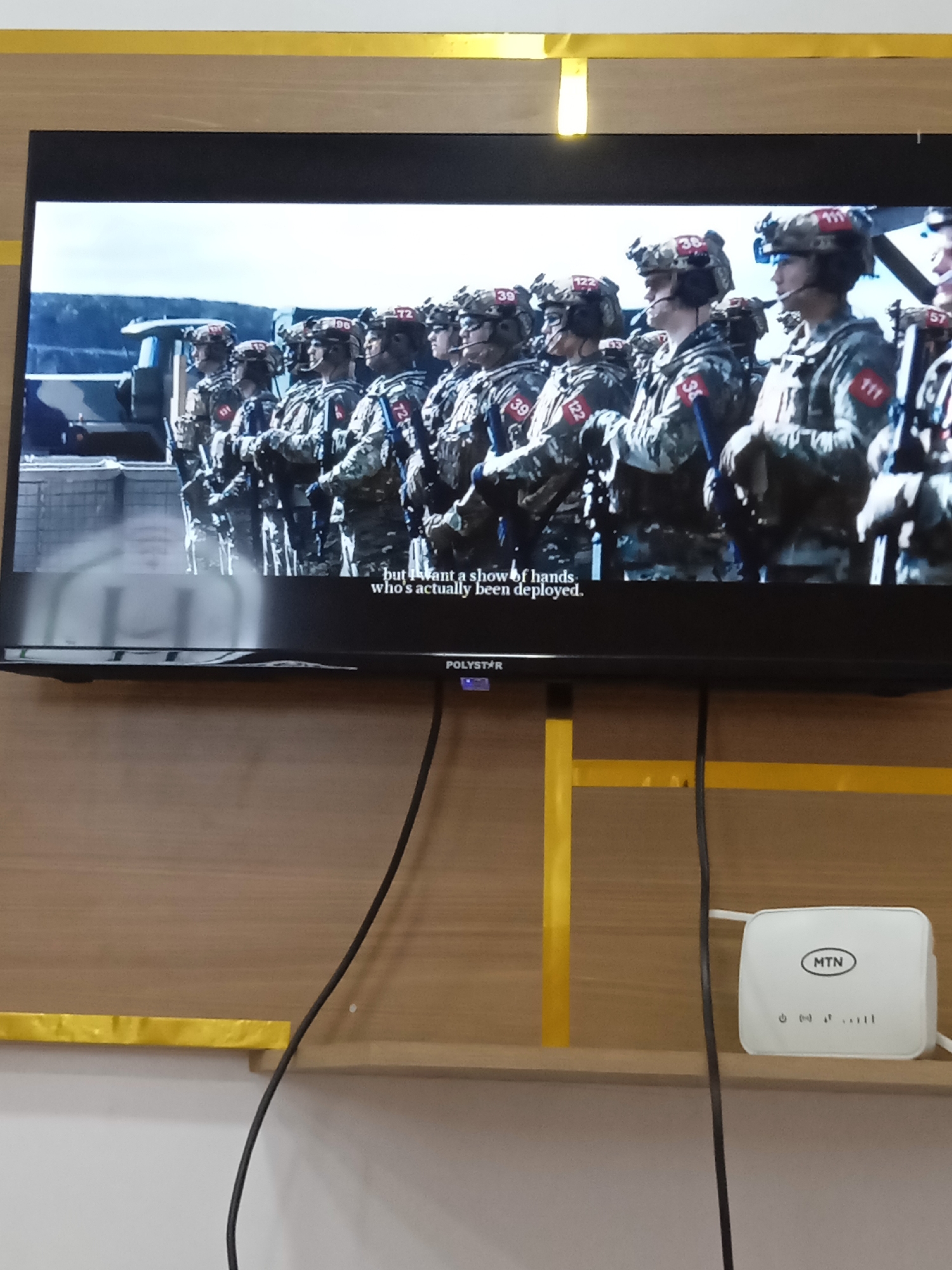 The image shows a television mounted on a wood-paneled wall, displaying a scene of soldiers in camouflage uniforms standing in formation. Each soldier has a number displayed on their helmet and uniform. Subtitles on the screen read, "but I want a show of hands who's actually been deployed." Below the television, on a shelf, sits a white MTN wireless router. The location is unknown.
