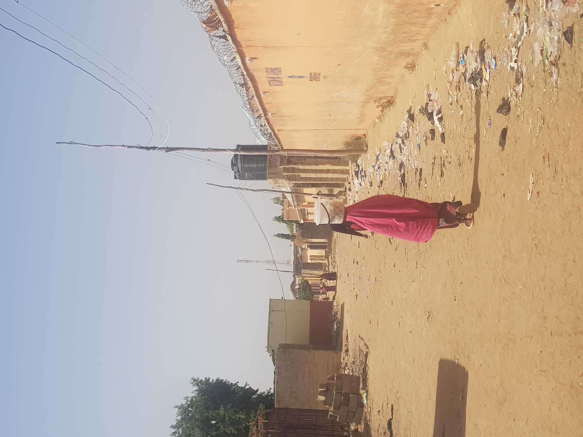 A sunlit, outdoor scene in Dabawa, Nigeria. A person in a red garment is walking on a dirt road. Litter lines the roadside next to a tall, peach-colored wall topped with barbed wire. Utility poles and wires traverse the sky. A water tank is situated on a building near the barbed wire. Buildings, bricks, and vegetation can be seen in the background. The sky is clear and pale blue.
