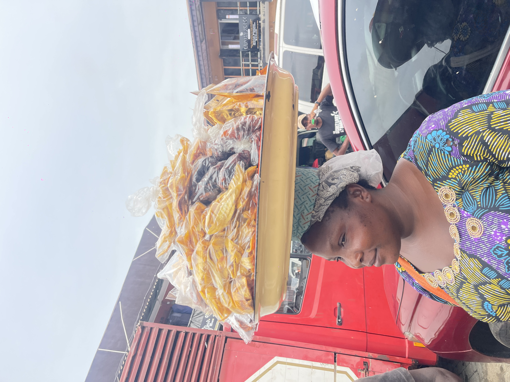 The image captures an outdoor street scene during the daytime, under an overcast sky, likely in an African country. The main subject is a woman positioned on the right side of the frame, looking towards the left with a calm, slightly smiling expression. She wears a patterned headwrap and a vibrant, colorful top featuring geometric designs in purple, yellow, blue, and black, adorned with circular, gold-colored embellishments around the neckline.
On her head, she expertly balances a large, light yellow rectangular tray laden with numerous clear plastic bags. These bags are filled with what appear to be snacks or dried food items; some are golden-yellow, resembling plantain chips or dried fruit, while others are darker in color, possibly nuts or different dried goods.
Behind the woman, to her left, is the side of a large red truck or bus. Further back, a red car is partially visible, and in its reflection, a man wearing a dark grey t-shirt with the text "BESTONES GANG" can be seen. In the background, buildings line the street, suggesting a commercial or market area. One building has a sign with partially visible text, which reads "LAPOSN PLACE". Another storefront sign, visible behind the red truck, appears to say "ORIGIN OF WEARS". The scene depicts the woman engaged in street vending, showcasing a common sight in bustling urban environments.