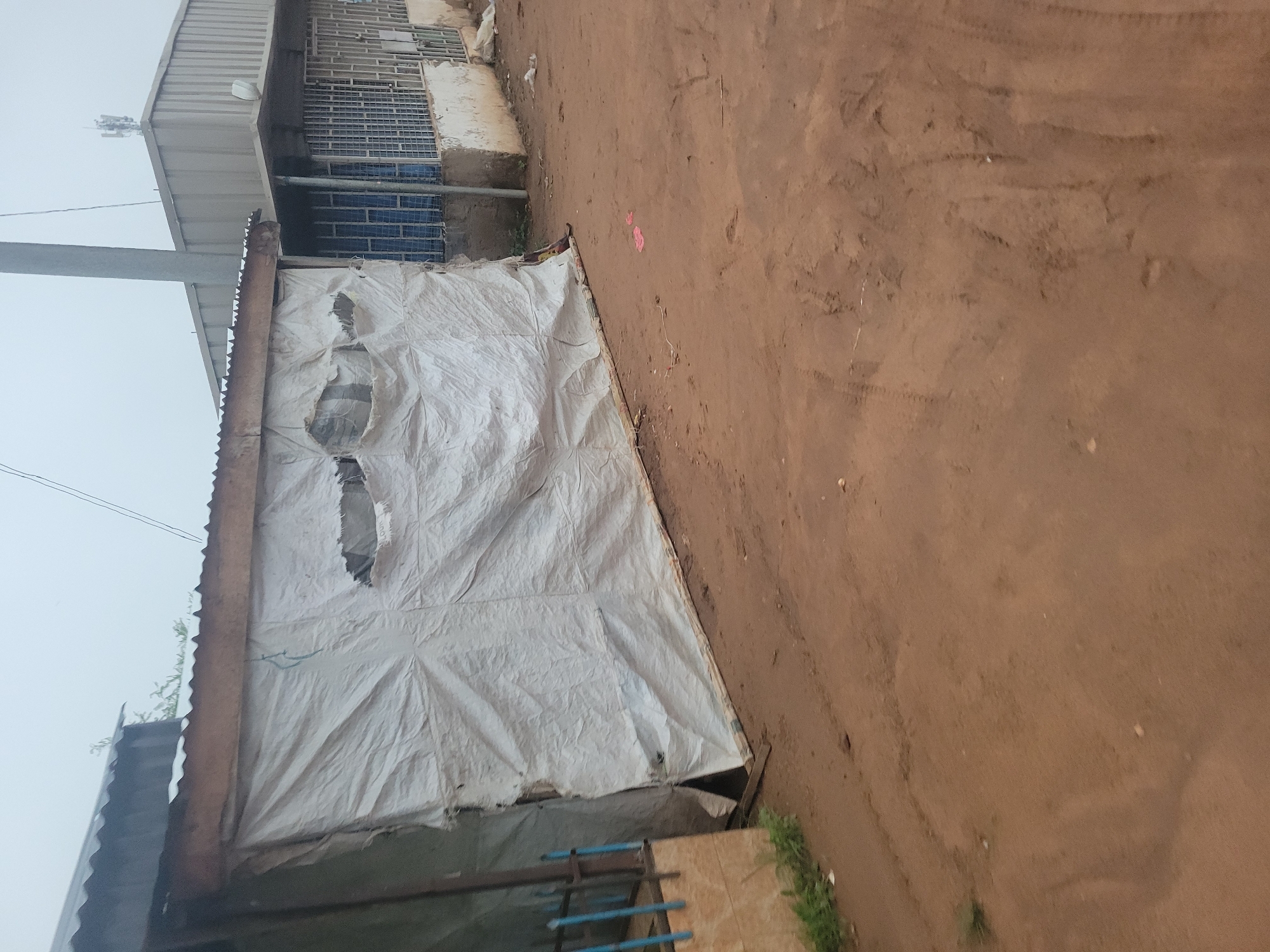 A temporary structure with a corrugated metal roof and white tarp walls stands on a dirt road. The tarp is torn in several places, revealing the interior. To the right, a more permanent building with a corrugated metal roof and barred windows is visible. The environment appears to be a roadside in an urban or semi-urban area, possibly with some light industrial or commercial activity. The sky is overcast.