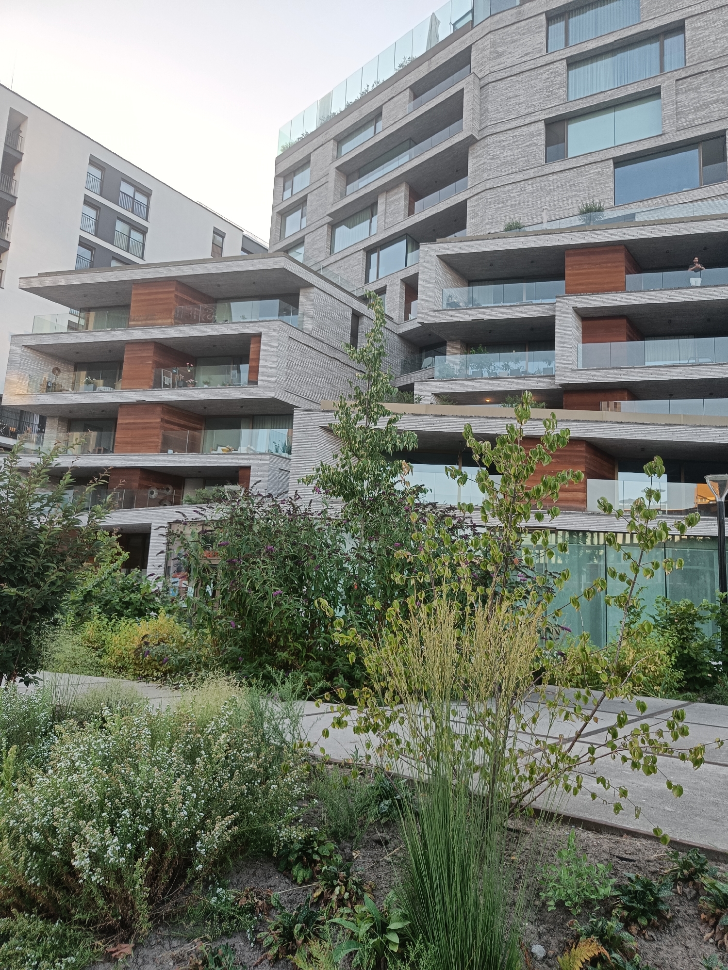 The image captures a modern building complex with a tiered facade, featuring balconies and large windows, in Leuven, Belgium. The building is constructed with light-colored brick and accented with wooden elements and glass railings. In the foreground, a landscaped garden area with various plants, including flowering bushes and ornamental grasses, is visible. A person is standing on one of the balconies, looking out. The sky is overcast and bright, suggesting it is daytime. The architecture indicates a residential or office building.