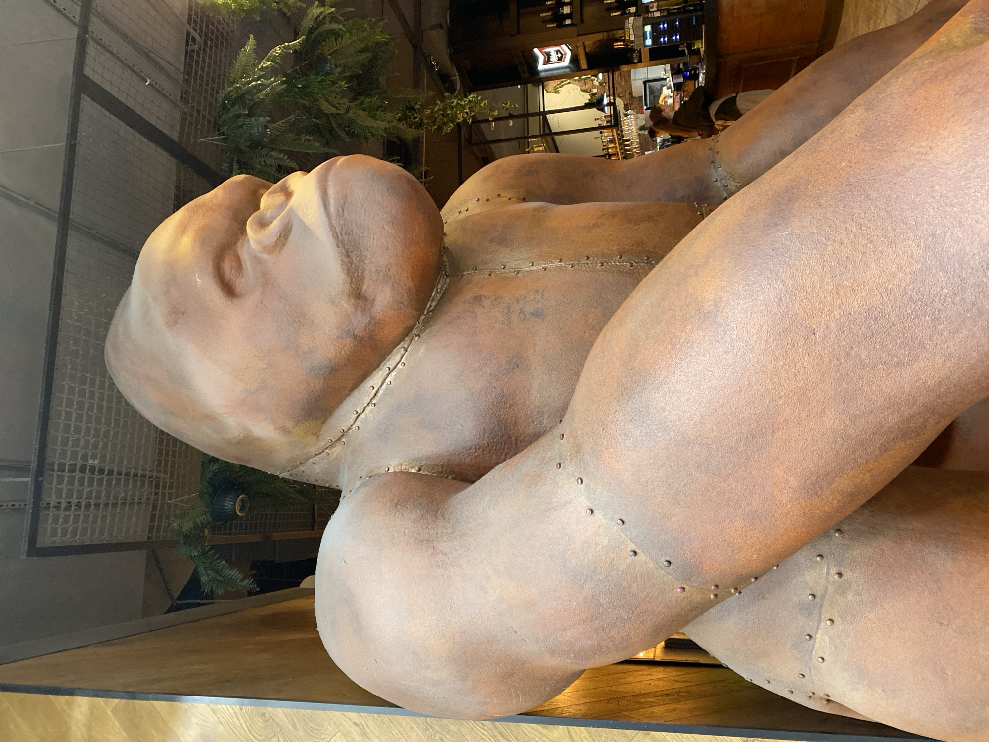 The image shows a close-up of a large, abstract sculpture of a reclining figure in a semi-industrial style. The sculpture is predominantly a rusty brown color with visible seam lines and rivet-like details, suggesting a constructed or fabricated quality. The figure is composed of large, rounded forms that resemble a human head and limbs, but are not anatomically precise. The head is tilted back with the eyes closed, conveying a sense of rest or contemplation.
In the background, glimpses of an interior space are visible, including a bar area with bottles and lighting fixtures. There are also some decorative green plants. The statue is located in the restaurant "Rachesy" in Yekaterinburg, Russia. The surroundings appear to be modern and stylish, suggesting the sculpture is an artistic element within the restaurant's decor.