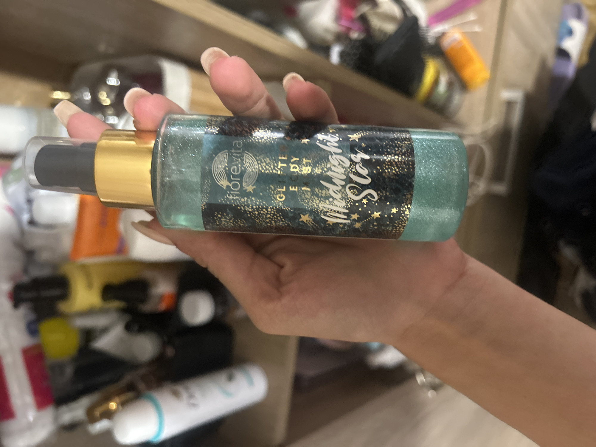 A person is holding a bottle of "fiorevita Glitter Body Mist Midnight Star" in front of a shelf filled with various personal care products. The bottle is partially filled with a shimmering turquoise liquid. The person's hand, with long acrylic nails, is visible holding the bottle. The background shows a cluttered shelf with items like a white spray bottle, a yellow bottle, a black bottle with an orange lid, and other unidentifiable products. The overall environment appears to be a bathroom or vanity area. The image does not provide any information about the city of Perushtitsa, Bulgaria.