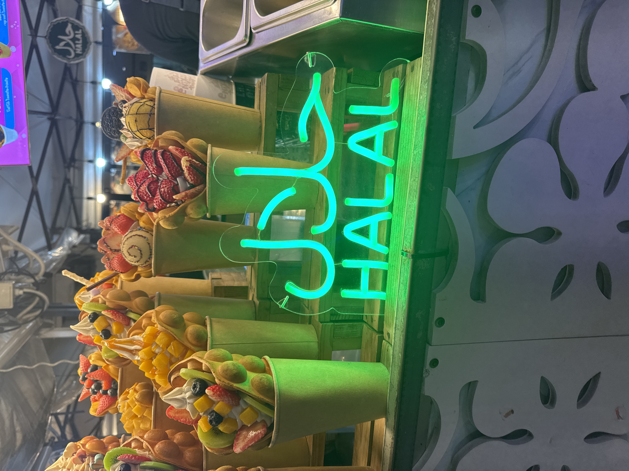 The image shows a food stall in Karon, Thailand, specializing in bubble waffles topped with fruits and ice cream. In the foreground, a bright green neon sign displays "HALAL" in both Arabic script and English. Several bubble waffles in green and tan paper cups are arranged on wooden shelves. The waffles are filled with a variety of colorful toppings, including strawberries, blueberries, kiwi, mango, and whipped cream. In the background, a person wearing a black shirt is visible, and the stall appears to be under a tent. The overall scene depicts a vibrant street food vendor.