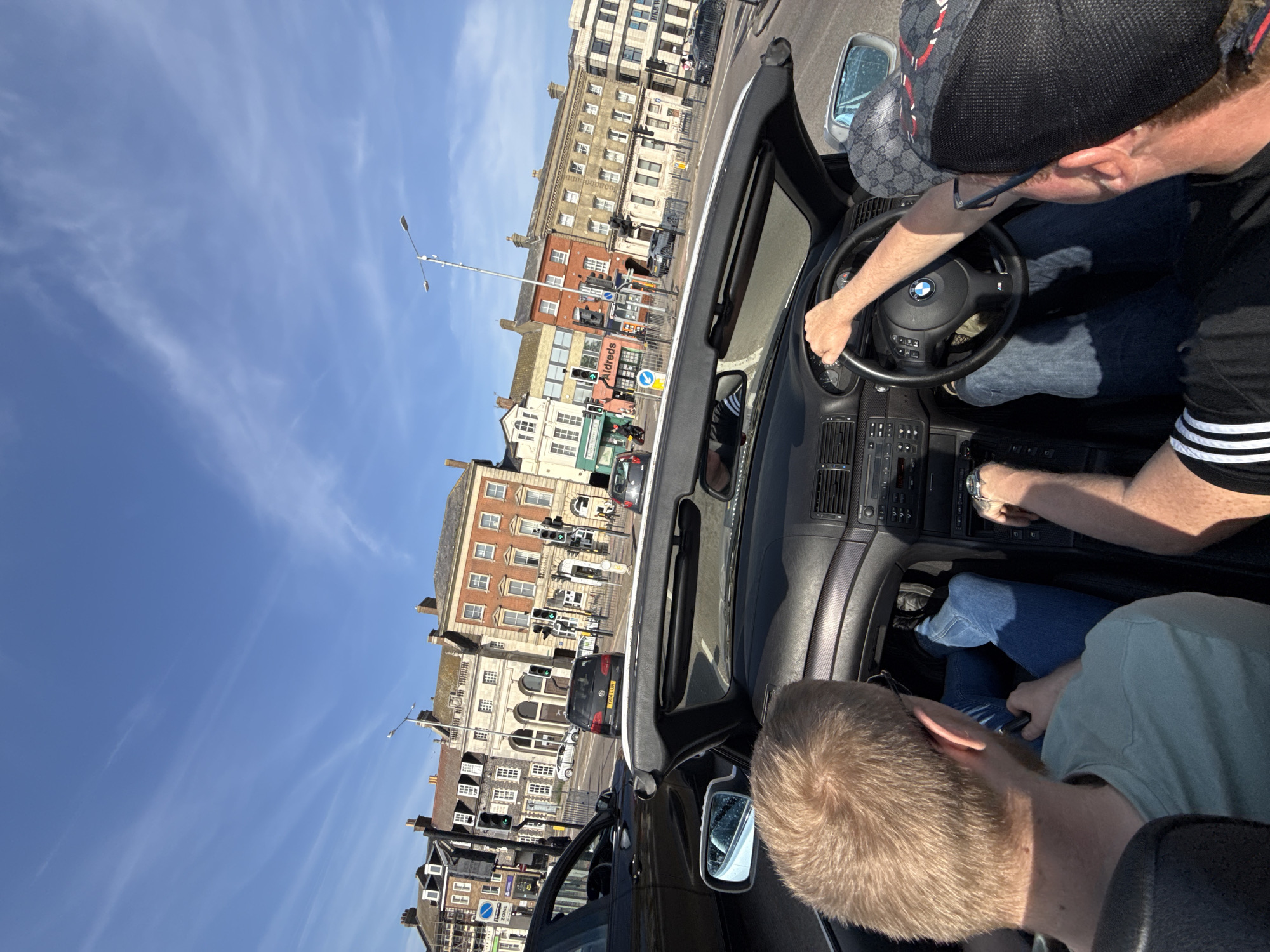 The image shows the interior of a black convertible car, likely a BMW based on the steering wheel logo, with the roof down, driving through Great Yarmouth, United Kingdom. The car is occupied by two men. The driver, wearing a black baseball cap with a Gucci pattern, is holding the steering wheel. The passenger, who is also wearing a pale green t-shirt, is looking down towards his phone. The buildings visible outside the car window reveal the urban environment. A traffic light is visible and street signs. There's a clear blue sky in the background.