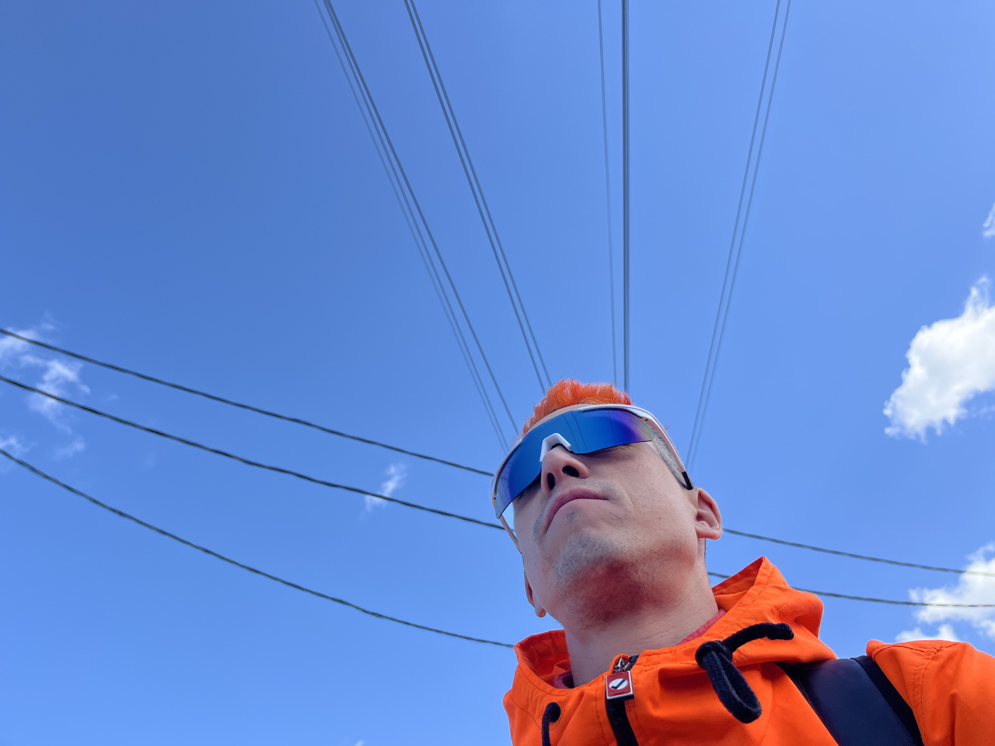 A low-angle shot depicts a person with orange hair wearing blue sunglasses and an orange jacket, possibly in the city of Chaikovsky, Russia. Overhead power lines stretch across the clear blue sky, punctuated by sparse white clouds. The person appears to be looking upwards. A black backpack strap is visible on their shoulder.