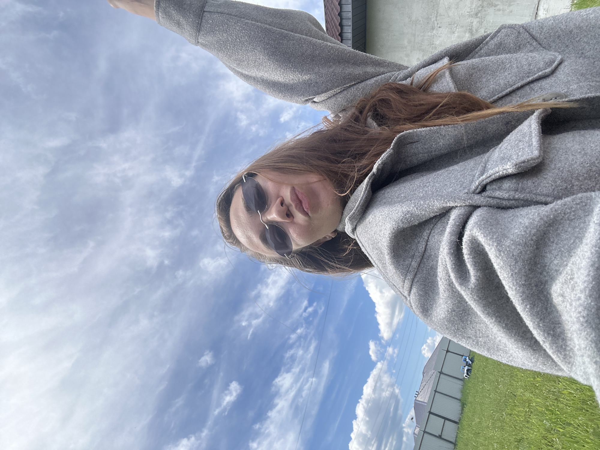 An outdoor selfie captures a person, likely female, with long, reddish-brown hair, wearing dark sunglasses and a grey, textured coat or jacket. Her head is tilted slightly, and she looks directly at the camera with a neutral expression.
The background is dominated by a bright blue sky dotted with scattered white, wispy clouds, suggesting fair daytime weather. Below the sky, to the right of the subject, a patch of vibrant green grass is visible. Behind the grass, a sturdy grey fence separates the foreground from what appears to be a residential area. Parts of a building, possibly a house with a dark tiled roof and a light-colored wall, can be seen beyond the fence. Two small white objects, resembling toy vehicles, are placed on the grass near the fence.
The photograph is taken from a low angle, with the person's arm extended upwards to capture the shot, encompassing a broad view of the sky and the outdoor environment. The setting appears to be a residential or suburban landscape, consistent with the given location context of Kharnavy, Russia. There is no visible text in the image.