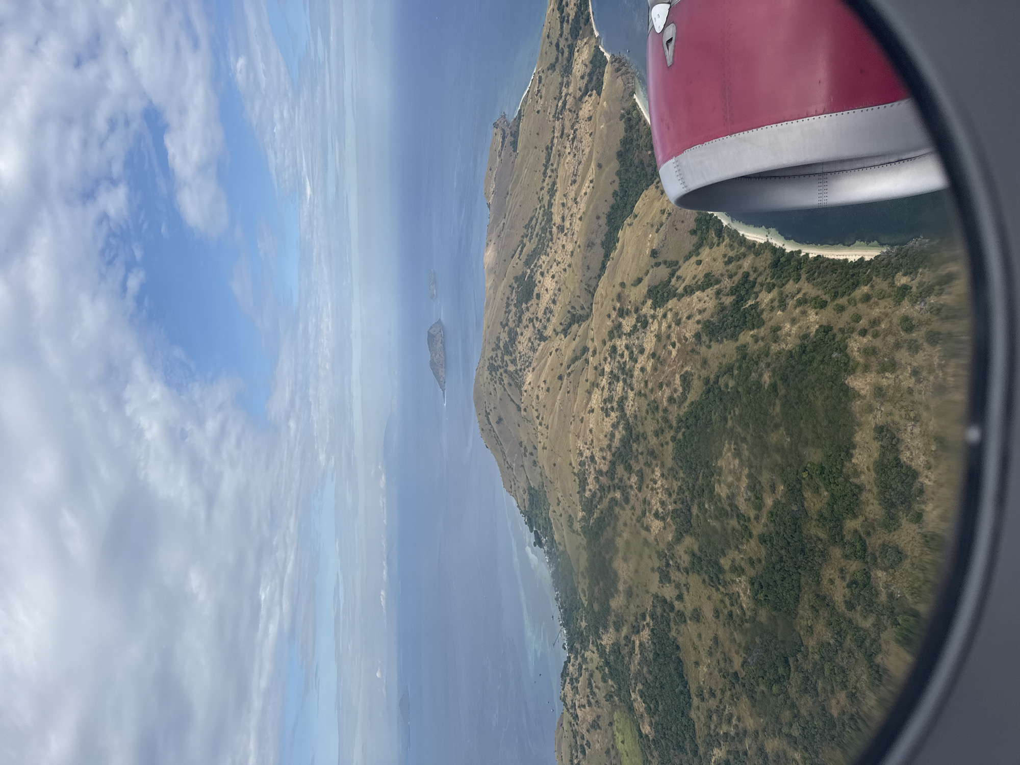 The image shows a view from an airplane window. Part of the plane's wing is visible in the upper right. Below the wing, there's land covered in vegetation. To the left of the land, there is a large body of water with a small island in the distance. The sky is visible in the upper left, with scattered clouds.