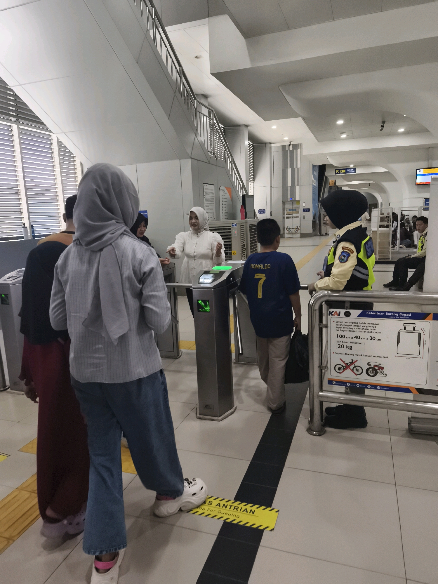 In the city of Palembang, specifically in Kelurahan 16 Ilir, Kecamatan Ilir Timur I, Indonesia, at what appears to be a train station or a similar transit point, a group of people are navigating through an electronic gate system. In the foreground, a person wearing a grey hijab and a striped grey long-sleeved shirt is seen from behind, with a maroon-colored skirt or dress visible on the person to their left. To the right of the gate, a uniformed individual, likely a security guard, is standing and observing. This guard is wearing a dark hijab and a yellow reflective vest over a lighter colored jacket, with patches on the vest indicating their affiliation. A young boy wearing a blue t-shirt with "RONALDO 7" on the back is walking through the gate. Behind him, another person in a white hijab and white outfit appears to be interacting with someone or operating the gate. The environment is modern, with white tiled flooring, metal railings, and signs in Indonesian, including one indicating luggage restrictions. The overall scene depicts orderly movement through a controlled entry point in a public transportation hub.