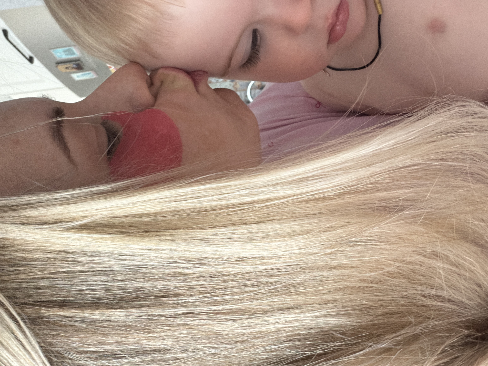 A close-up, skewed-angle shot depicts a fair-skinned child with blonde hair resting their head on a light-skinned adult with blonde hair. The adult's eyes are closed, and pink patches or stickers are applied under the eyes. The child's eyes are mostly closed with visible eyelashes. The child is wearing a thin black necklace with gold accents. The background is blurred, but hints of a kitchen or refrigerator are visible. The exact location in the city of Votkinsk, Russia, cannot be determined from the image.