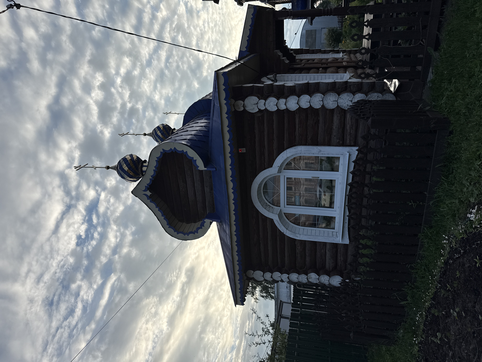 The image shows a small, wooden Orthodox church with blue onion domes, located in the village of Kukuy, Russia. The church is constructed of dark logs, with decorative white trim around the roofline and a distinctively shaped entryway. A white-framed arched window is visible on the side of the building, reflecting an outdoor scene. The church is surrounded by a dark, wooden picket fence and lush green grass. In the background, a metal fence and some vegetation can be seen, along with a cloudy sky. There are no people or animals visible in the image. The scene appears to be captured during daylight hours.