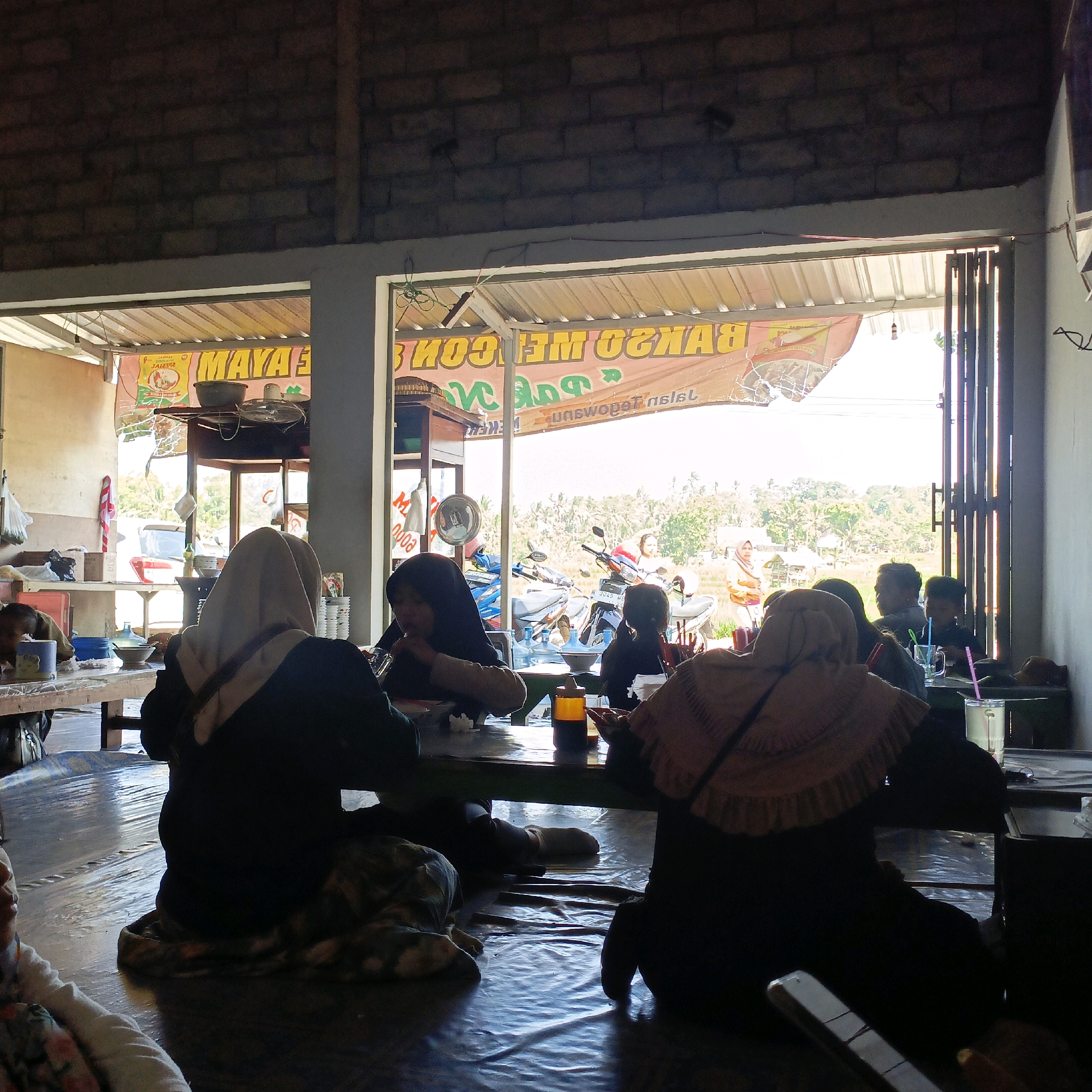 The image depicts the interior of a modest, open-air eatery in Salatiga, Indonesia, with patrons seated at tables on a mat-covered floor. The eatery appears to be a roadside establishment, possibly a "warung" or simple restaurant, with a corrugated metal roof extending over the seating area. Banners with Indonesian text, including "MAYA" and "BAKSO ME GOM," are visible on the exterior, suggesting the establishment's offerings.
In the foreground, several individuals are seen from behind, engaged in eating or conversing. They are dressed in casual attire, with some women wearing hijabs. The tables are simple, wooden structures, some covered with green cloths. On the tables, there are bowls, glasses with straws, a bottle of seasoning, and other dining implements. The floor is covered with a plastic mat, which has some ripples and creases, indicating it might be a temporary or protective covering.
Behind the seated patrons, through an open doorway and windows, a sunny outdoor scene is visible. This includes a dirt or gravel area where motorcycles are parked, and beyond that, a rural landscape with rice paddies and vegetation under a bright sky. A few people are seen standing or walking in the background outside. The architecture of the building is rudimentary, with exposed brick walls and metal supports. The overall atmosphere is informal and functional, typical of a local eatery in a less urbanized setting in Indonesia.