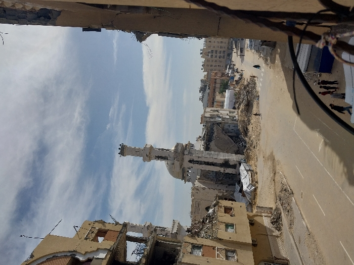 This image depicts a scene of widespread structural devastation in Khan Yunis, Palestinian Territory, under a partly cloudy sky. The foreground reveals a partial view of a damaged structure on the right, with dark metal elements. A paved street with white lane markings runs through the center, containing scattered debris.
On the left, a multi-story building exhibits severe damage, with large sections of its facade collapsed, exposing internal rooms and significant rubble at its base. Dominating the midground is a severely damaged mosque, characterized by a tall, ornate minaret that remains largely intact, and a partially collapsed dome. The mosque's main structure shows extensive destruction, with considerable debris and rubble accumulated around its foundation and extending into the street. White tarp-like material is visible amidst the rubble near the mosque.
Several individuals are present, including a group of approximately 5-7 people, mostly men or boys, gathered near a small blue and white structure in the foreground. Other individuals, including one in dark, long clothing, walk on the street and through the debris in the midground. The background shows additional damaged urban buildings and what appears to be a large white tent structure further down the street. The environment is characterized by extensive destruction and debris.