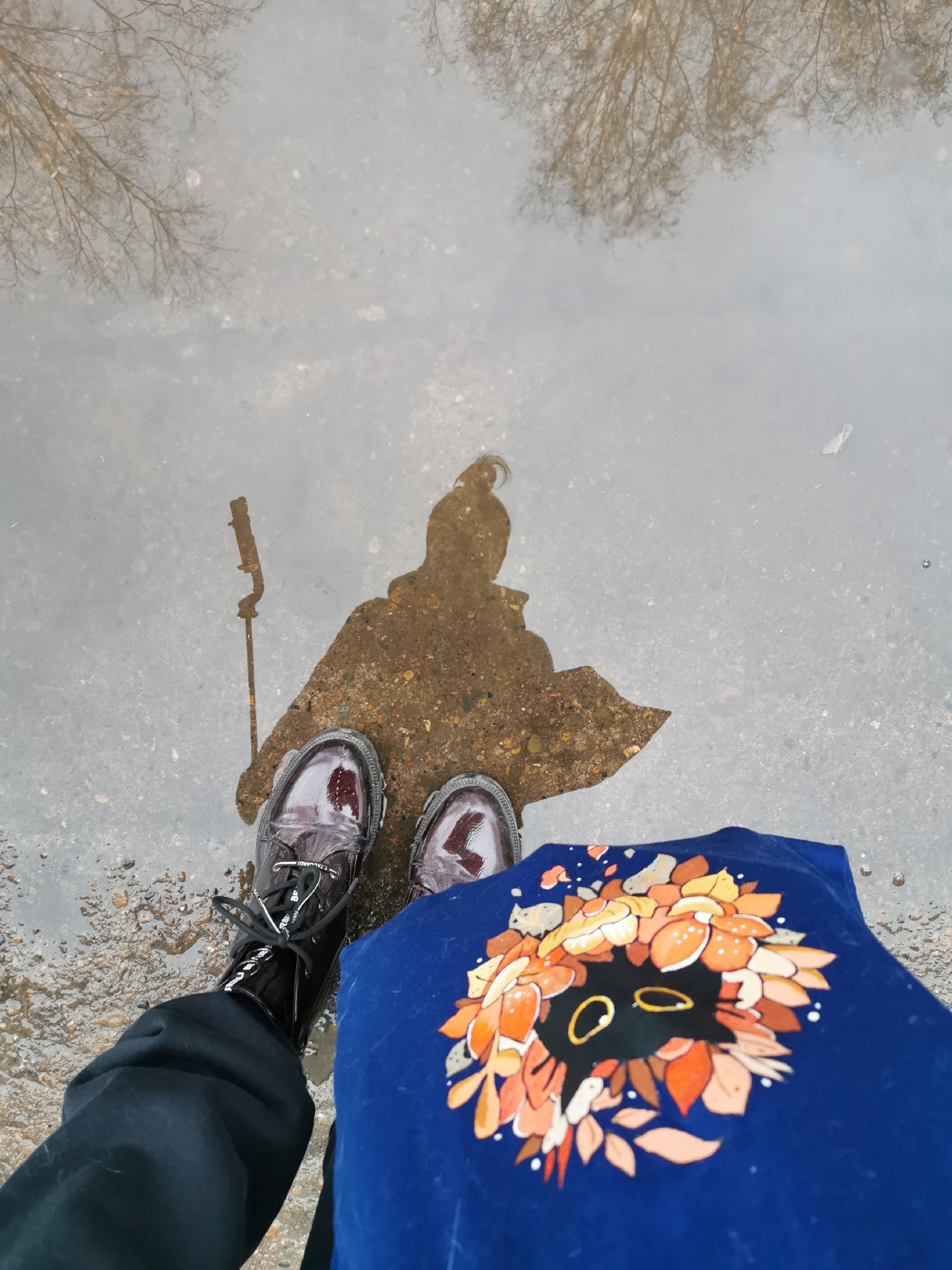 The image shows a first-person view of someone standing in a shallow puddle on a paved surface. The puddle reflects the person's silhouette, a stick-like object, and bare trees. The person is wearing shiny dark boots and dark pants. A blue t-shirt with a design of leaves surrounding a black cat face is partially visible in the lower right corner of the image. The location appears to be an outdoor urban area.