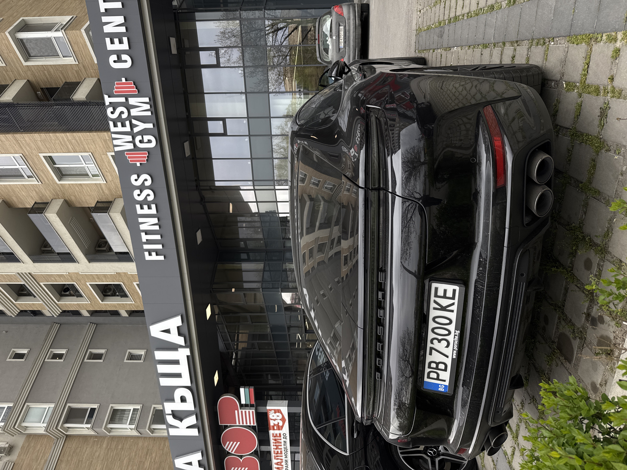 The image shows a black Porsche parked in front of a building complex in Plovdiv, Bulgaria. The vehicle's license plate is clearly visible, reading "PB 7300 KE" and the country abbreviation "BG" for Bulgaria. Part of a gray car is visible in the background. The building behind the car houses a "Fitness West Gym Center" and a store with a prominent sign indicating a "38% discount on selected models". The structure's architecture features modern elements, including glass panels and beige facades. Paving stones are visible.