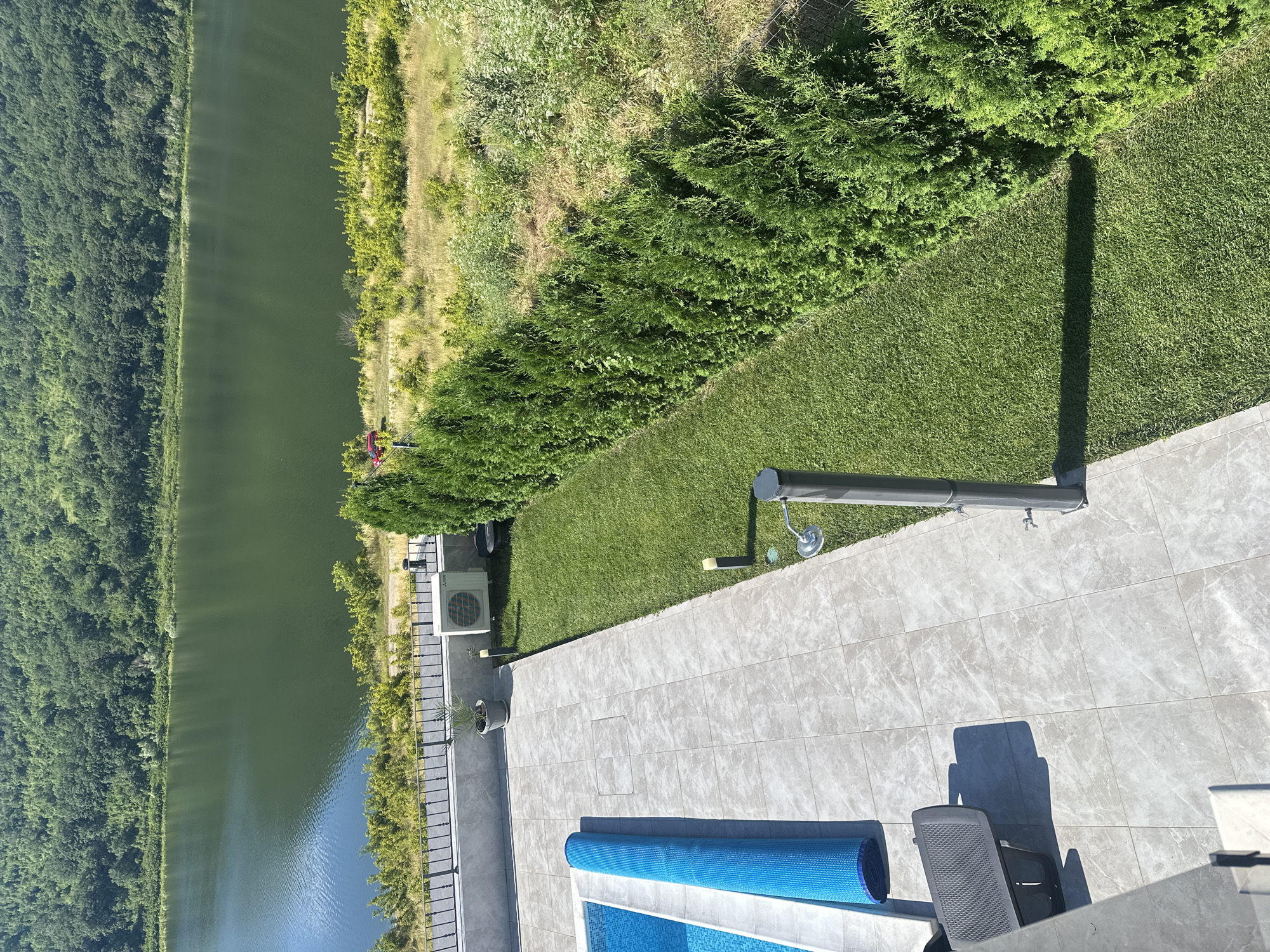 An aerial shot captures a serene lakeside view with a lush green forest backdrop. In the foreground, a pristine swimming pool is partially covered with a blue rolled-up cover, adjacent to a tiled patio. A grey lounge chair sits on the patio, casting a shadow. A modern outdoor shower stands near the edge of the lawn, which is bordered by a neatly trimmed hedge. An air conditioning unit is visible on the tiled area. The lake reflects the clear blue sky, and beyond it, the dense forest stretches towards the horizon, suggesting a rural or natural setting.