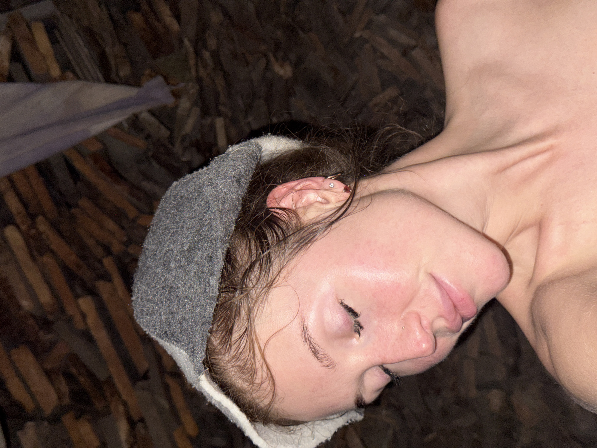 The image shows a close-up of a woman's face and shoulders. She is wearing a gray cap with a white trim and has her eyes closed, with her head tilted down. Her hair is wet and appears to be pulled back. She has light skin and appears to be in a sauna or steam room, indicated by the steamy appearance of her skin and the rustic wooden background. There are no visible landmarks or indications of the city of Maksimovo, Russia, in the image.