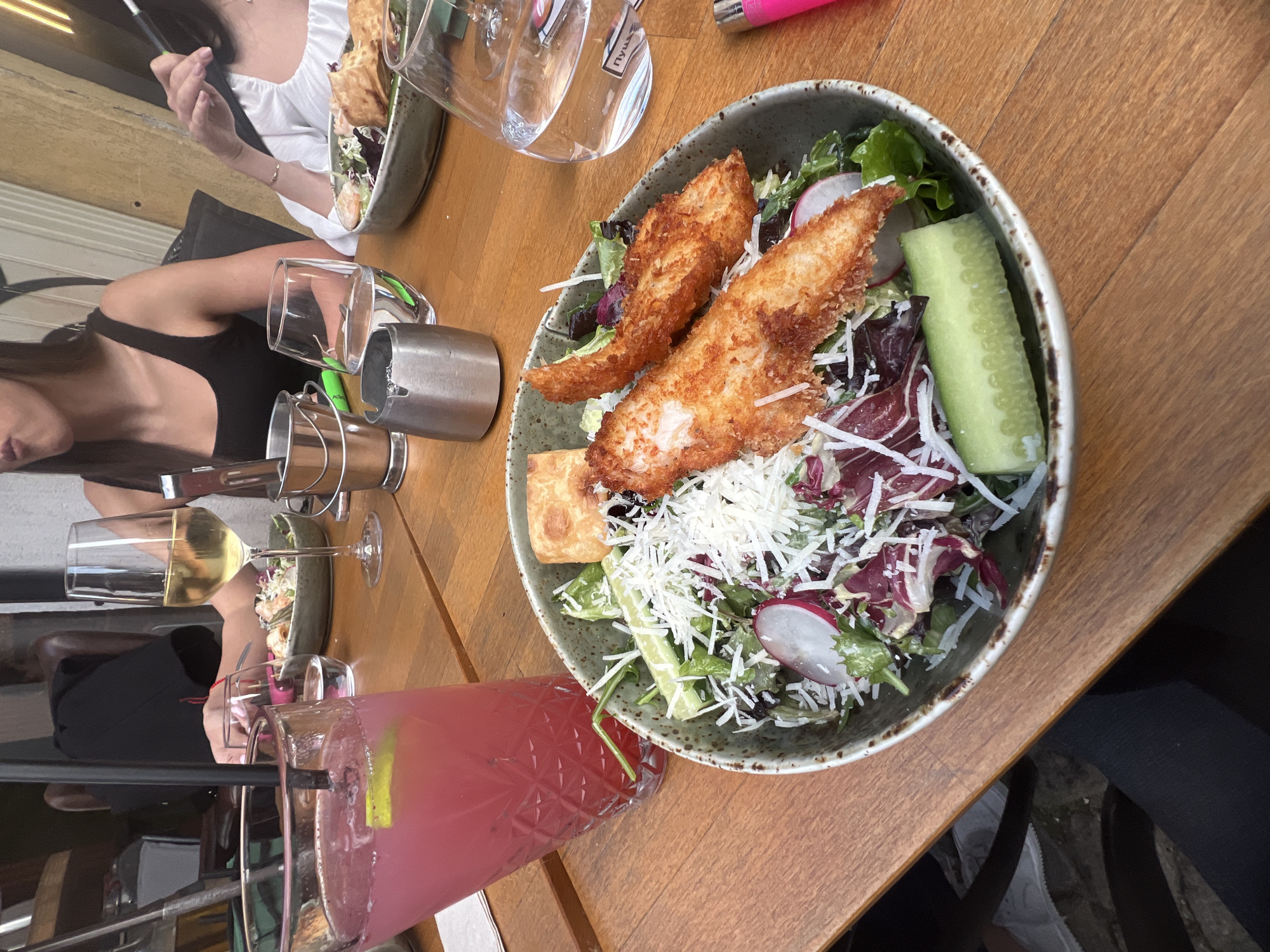 The image depicts a casual outdoor dining scene at a restaurant or cafe in Plovdiv, Bulgaria, during bright daylight. A central focus is a large, textured bowl filled with a fresh salad. The salad comprises mixed green and red lettuce, sliced radishes, a thick cut of cucumber, and a generous topping of shredded white cheese. Two pieces of golden-brown, breaded and fried protein, likely fish or chicken, are prominently placed on the salad, alongside a small piece of flatbread or cracker.
Several beverages are arranged on the light brown wooden table. In the foreground, a tall glass holds a vibrant pink cocktail, garnished with a lime wedge. To the left, a wine glass contains white wine, and two clear water glasses are also present; one has faint Cyrillic text visible, possibly "Пуш". Two women are partially visible: one in the upper left wears a white top and holds a dark, slim object (potentially a vape pen or hookah hose), while another in the middle-left wears a black tank top. Additional objects include stainless steel cylindrical holders for cutlery or napkins and a pink lighter. The overall scene conveys a relaxed social gathering enjoying a meal and drinks outdoors.