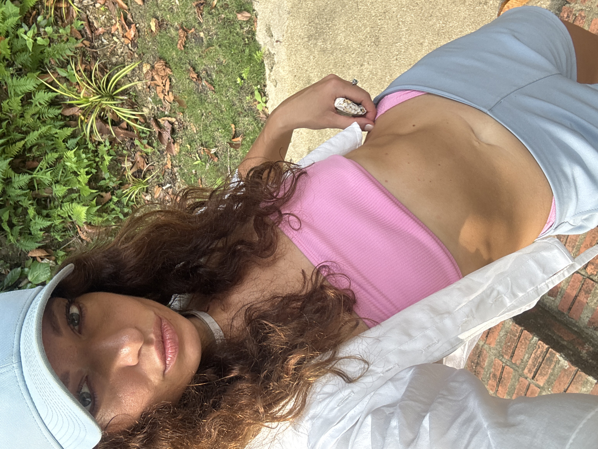 A woman wearing a light blue baseball cap, a pink top, and blue shorts poses for a selfie outdoors. She is positioned on a surface, possibly a bench, covered by a white shirt. Behind her are foliage, a patch of grass, and a sidewalk. A brick structure is also visible. No specific details suggest the location is Đà Nẵng, Vietnam.