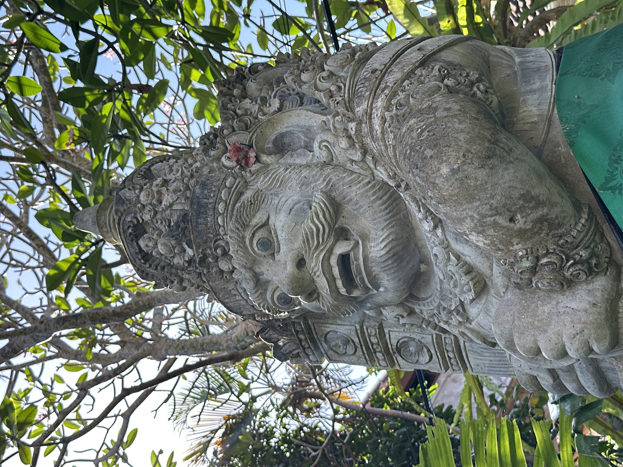 A close-up, low-angle shot captures a stone statue of a Balinese deity, likely in the Ubud District of Indonesia, against a backdrop of green foliage and a bright sky. The statue, weathered and detailed, depicts a fierce face with wide, staring eyes, a prominent snout, sharp teeth bared in a grimace, and a thick mustache and beard. It wears an ornate, multi-layered headdress and intricate jewelry on its arms and ears. A small red flower is tucked behind its right ear. The statue is adorned with a green cloth draped around its waist. Lush green leaves and tree branches fill the upper portion of the frame, with some dappled sunlight filtering through. The statue's massive hands are clasped in front of its body, grasping what appears to be a club or staff. The stone exhibits signs of age and exposure to the elements, with some moss and discoloration visible. The overall impression is one of imposing presence and ancient artistry, set within a natural, tropical environment.